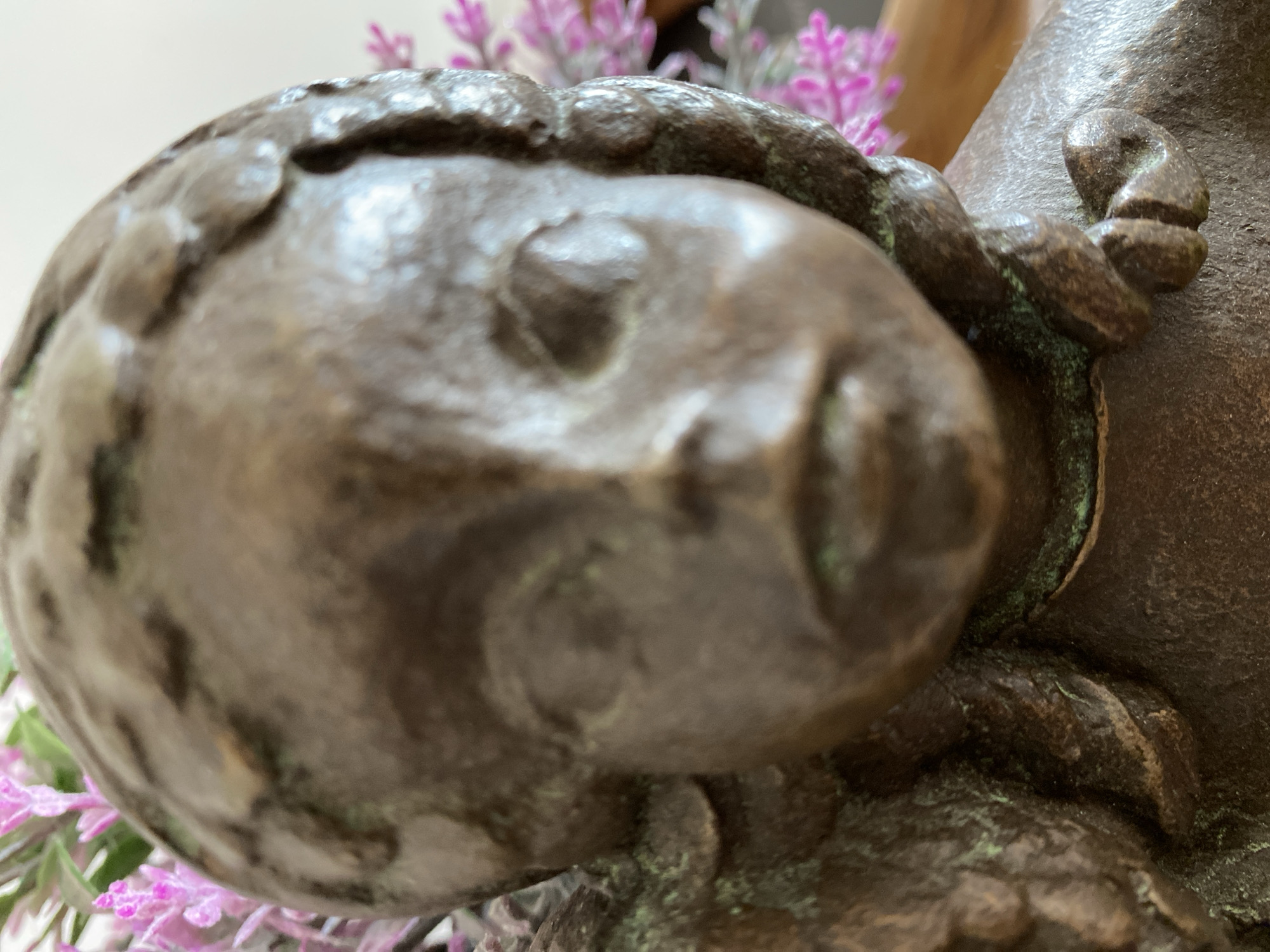 The image shows a close-up of a bronze statue of a child's face. The facial features are soft and rounded. The statue appears to be aged, with some green patina visible in the crevices. Behind the statue, there are blurry purple flowers. The exact location in Budva, Montenegro, cannot be determined from this image alone.
