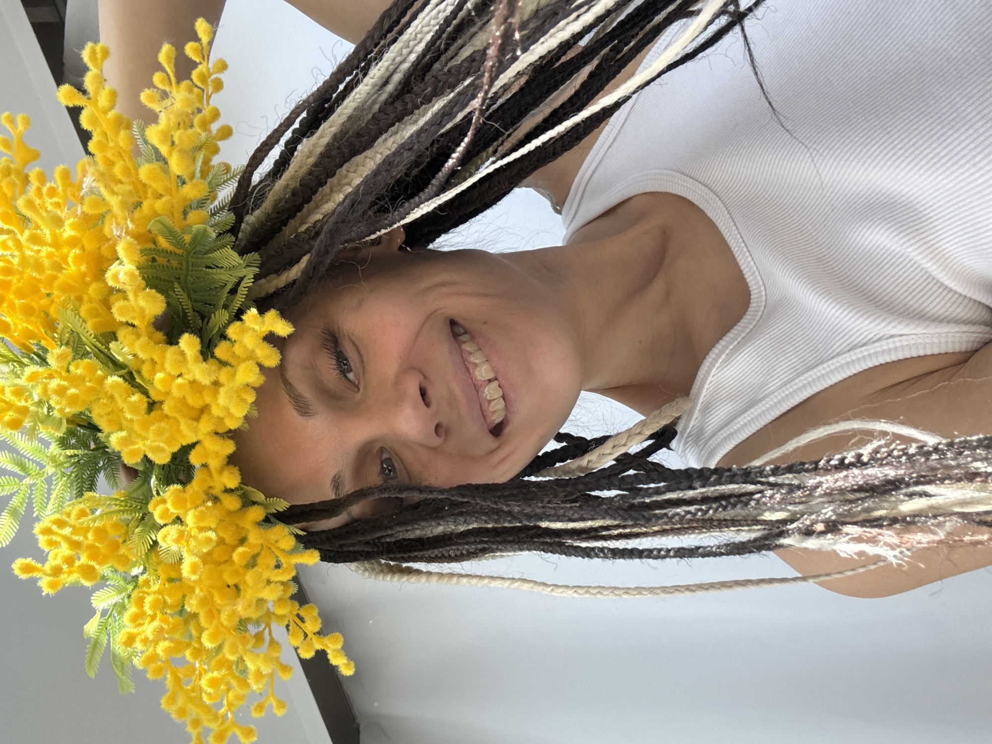 A portrait captures a young adult woman smiling broadly, presented from a low-angle perspective that makes her appear inverted or at a significant tilt. Her head is positioned towards the bottom of the frame, and she gazes directly at the viewer with a wide, genuine smile revealing her teeth, conveying happiness and contentment.
She has long dreadlocks or braids, featuring a striking mix of dark brown and lighter, possibly blonde or beige, strands that cascade around her face and extend upwards. Adorning the left side of her head is a large, vibrant yellow floral arrangement, resembling a crown or headdress. These fluffy, round blooms, characteristic of mimosa flowers, are interspersed with green faux leaves, adding a whimsical and celebratory touch. She is wearing a simple white ribbed tank top, which is visible on her torso extending towards the upper part of the image. The background is uniformly light and plain, suggesting an indoor setting or bright, diffused lighting. There are no discernible location cues or visible text in the image.