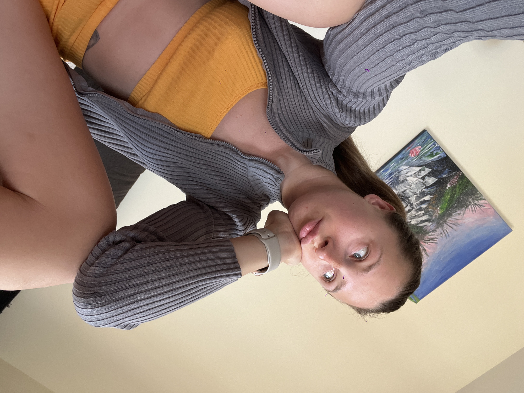 A woman is posing in front of a light-yellow wall with a framed landscape painting hanging on it. She wears a ribbed mustard-yellow top and a grey, ribbed zipper cardigan. She also has a watch on her wrist. A glimpse of her upper leg is visible on the left side of the image. The location, Bečići, Montenegro, is not evident from the image.