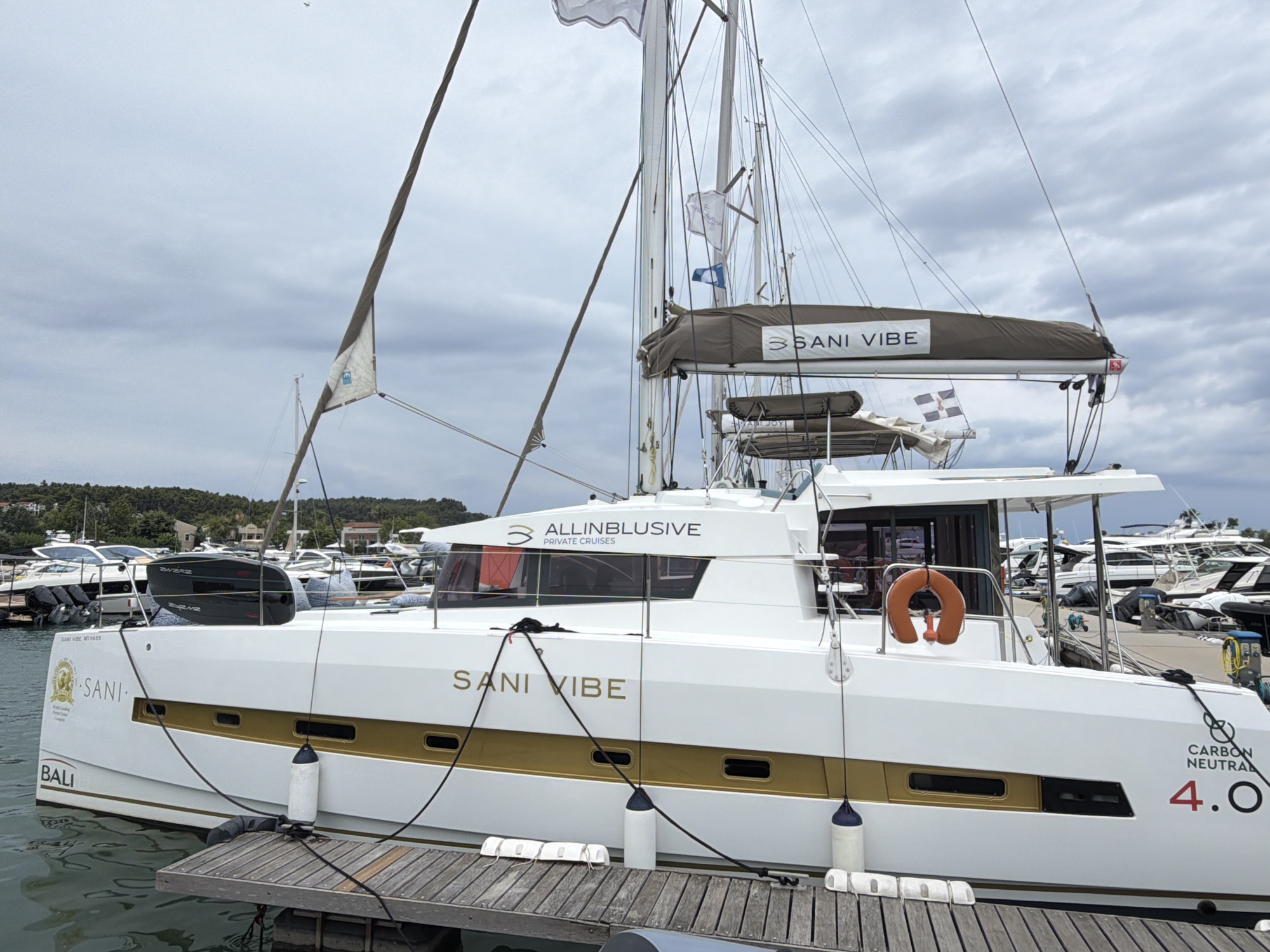 A white catamaran named "SANI VIBE" is docked at a marina, likely in Kassandra Municipal Unit, Greece, under an overcast sky. The catamaran has a gold stripe along its hull with several windows. The name "SANI VIBE" is printed in gold on the side of the hull. Above the windows, "ALLINBLUSIVE PRIVATE CRUISES" is written in dark blue text. A life preserver, orange in color, is attached to the railing on the starboard side. The boat is secured to a wooden dock with ropes. In the background, other boats and buildings are visible. The water appears calm. There are no people visible in the image.
