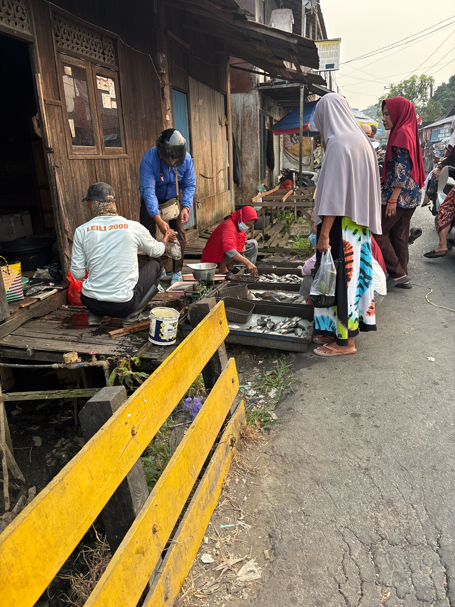This image captures a street scene in Banjarmasin, Indonesia, likely near a market or waterway, given the weathered wooden structures and the presence of fish for sale. In the foreground, a person wearing a blue jacket and a black helmet is interacting with another person who is crouching and wearing a white long-sleeved shirt with "LEILI 2000" printed on the back, and black pants and boots. They are on a wooden platform, with a canal or drainage ditch visible below. To the right, two women are engaged in activity. One, in a red headscarf and mask, is handling fish laid out on a tray, appearing to be a vendor. Another woman in a light-colored headscarf and a vibrant, patterned skirt is observing or interacting with the fish vendor. Behind them, more fish are displayed in trays, indicating a marketplace setting. The environment appears somewhat dilapidated, with aging wooden buildings and a paved road. The time of day seems to be late afternoon or early morning, judging by the quality of light. The overall impression is of a bustling, albeit humble, local commerce scene.