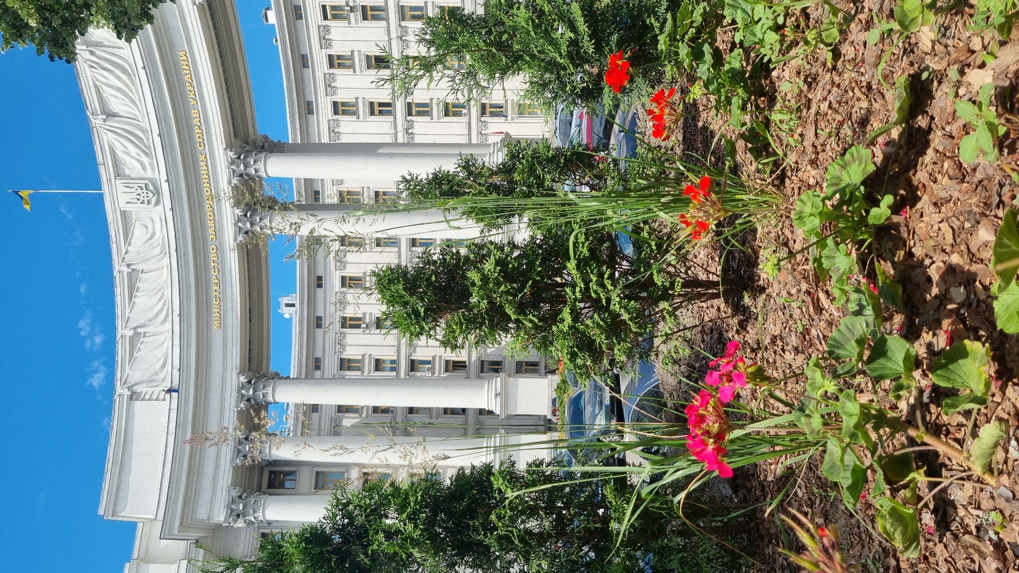 The image shows the Ministry of Foreign Affairs of Ukraine building in Kyiv, Ukraine. The building is a large, white, neoclassical structure with a curved facade, grand columns, and a prominent gold inscription that reads "МІНІСТЕРСТВО ЗАКОРДОННИХ СПРАВ УКРАЇНИ" (Ministry of Foreign Affairs of Ukraine). A Ukrainian flag flies from a flagpole on top of the building. In the foreground, there is a landscaped area with green coniferous trees, tall grasses, and blooming red and pink flowers. Several cars are parked in the background. The sky is a clear blue, indicating a sunny day.