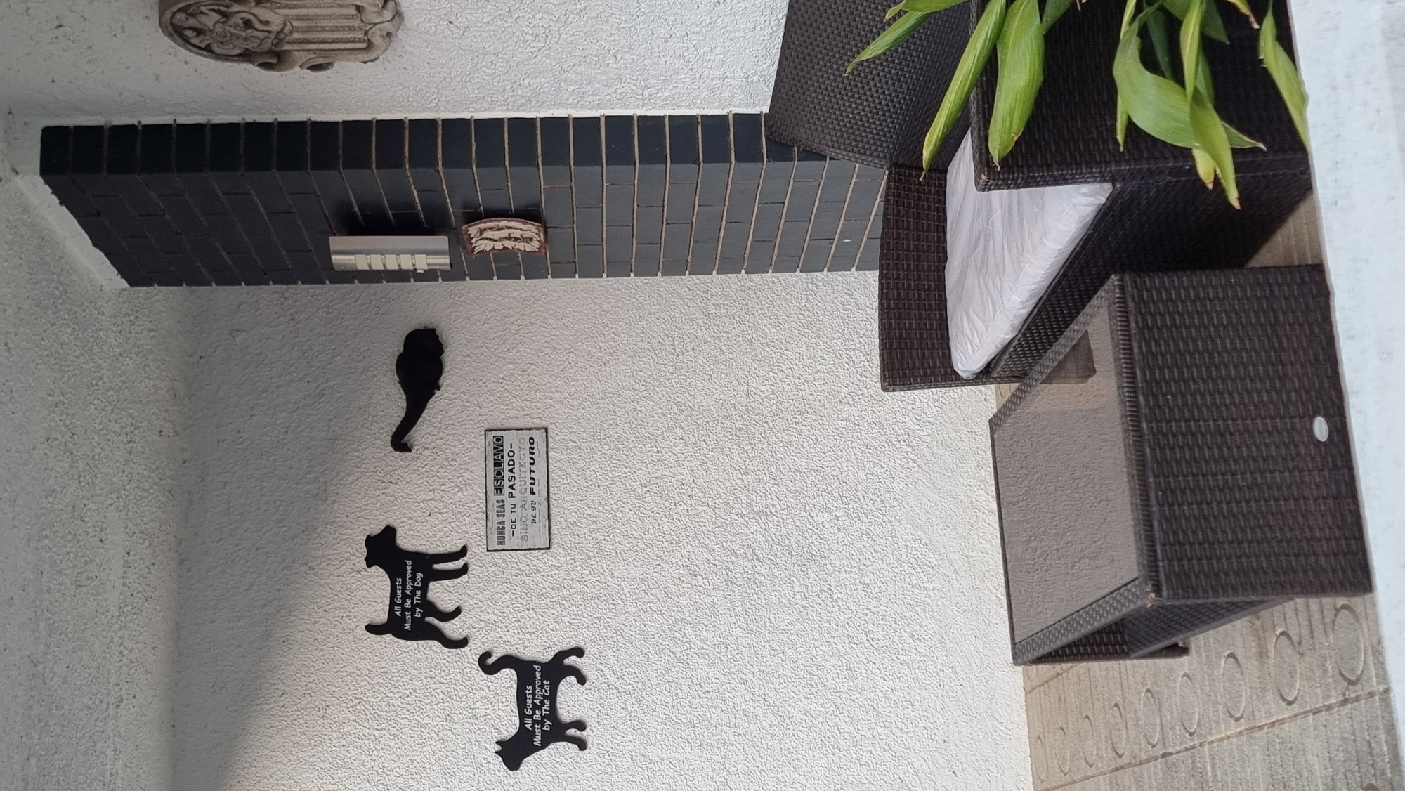 The image depicts an outdoor patio area with a textured white wall, a dark brick column, and a tiled floor. On the wall, there are several decorative black silhouette cutouts. One cutout is of a dog with the text "All Guests Must Be Approved by The Dog". Another is of a cat with similar text, "All Guests Must Be Approved By The Cat". There is also a silhouette of what appears to be a duck or bird. A small, framed sign with Spanish text hangs between the dog and bird silhouettes. The sign reads: "NUNCA SEAS ESCLAVO -DE TU PASADO- SINO ARQUITECTO DE TU FUTURO", which translates to "NEVER BE A SLAVE -TO YOUR PAST- BUT AN ARCHITECT OF YOUR FUTURE".
Adjacent to the wall, there is a dark wicker patio set consisting of a coffee table with a glass top and a sofa. The sofa has a white cushion. A large green plant is positioned next to the sofa. The overall scene suggests a relaxed, outdoor living space. There are no people visible in the image. The environment is that of a residential patio. Given that the city of Sant Joan Les Fonts, Spain is mentioned, this scene is likely located in a residential property within that municipality.