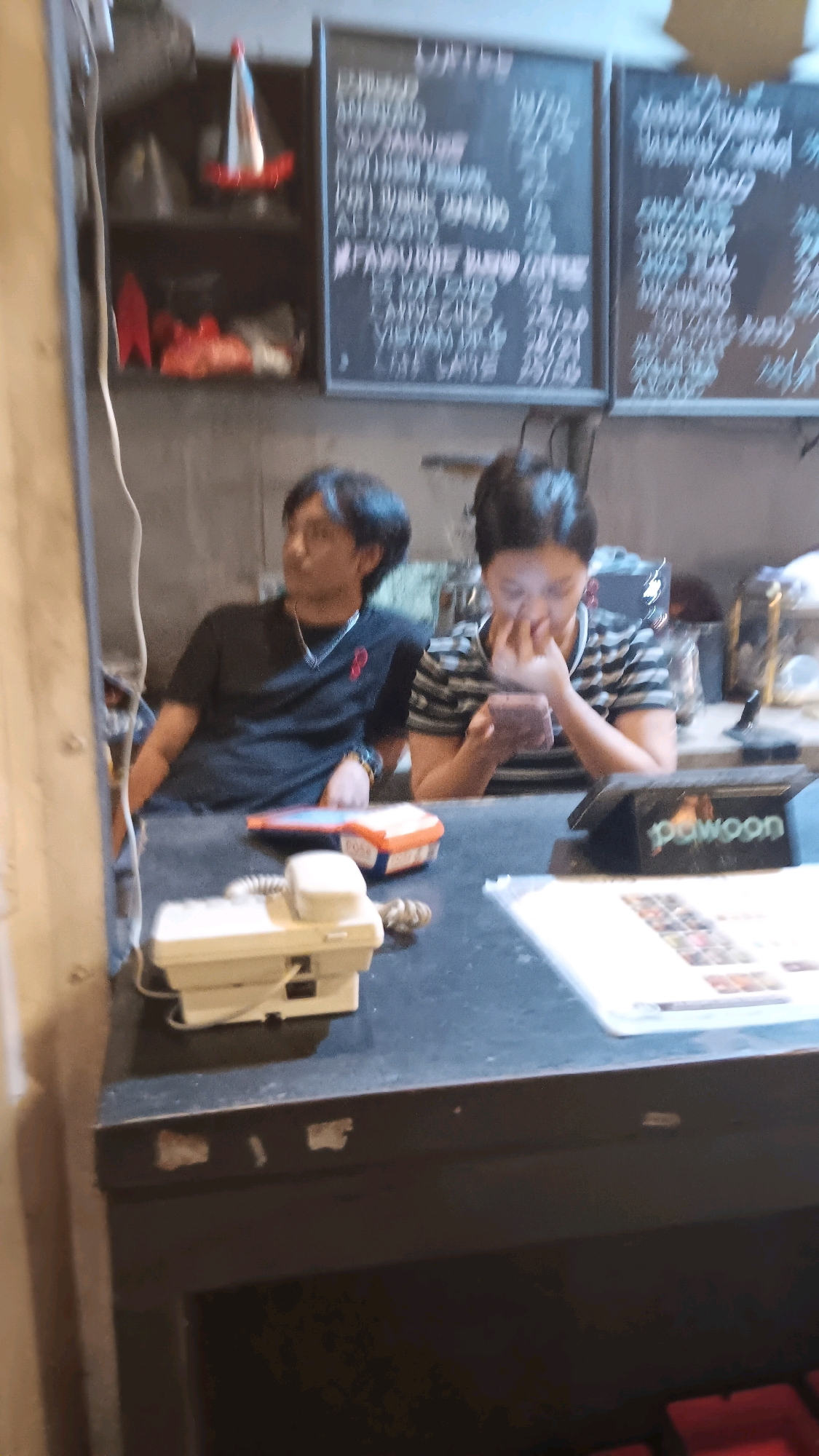 In a dimly lit establishment in South Jakarta, Indonesia, two individuals are seated behind a counter. On the left, a man with dark hair, wearing a black t-shirt with a red symbol on the collar and a silver chain, leans on his left arm. To his right, a woman with dark hair, wearing a black and white striped t-shirt, is engrossed in her mobile phone, her fingers touching her face. In front of them on the counter sits a white desk phone and a colorful container. Behind them, a menu board displays various coffee options.