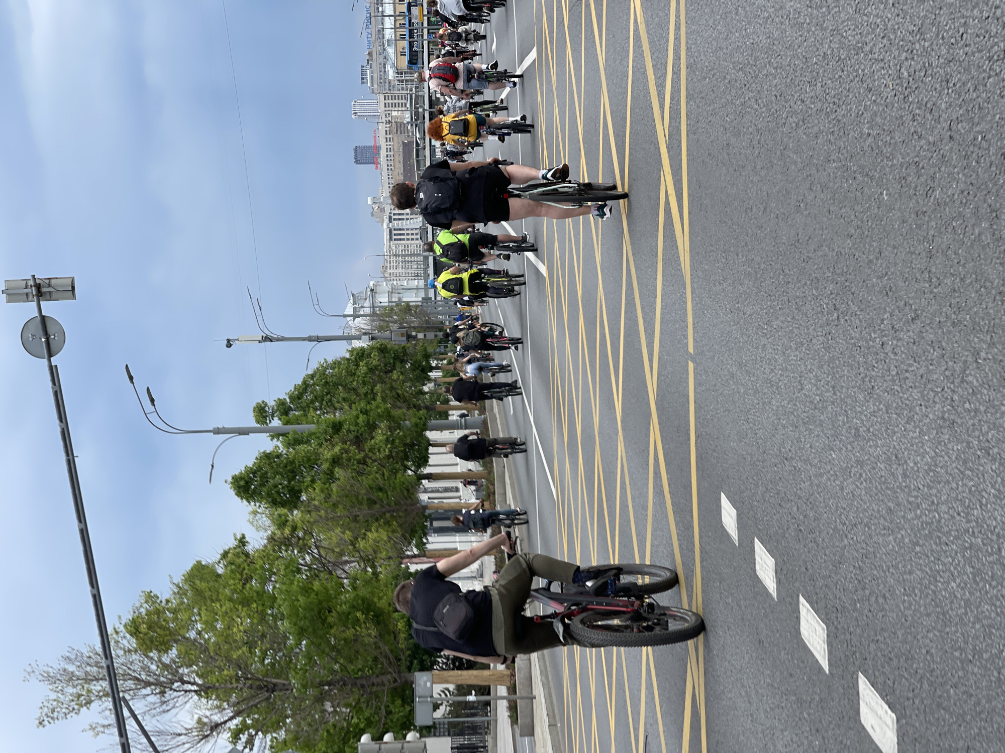 The image depicts a bicycle parade taking place on a street in Moscow, Russia. The scene is captured from the perspective of one of the cyclists, looking forward along the roadway. Several cyclists are visible ahead, riding on a gray asphalt road with a pattern of yellow crisscross lines and white dashed lines. Trees line the left side of the road, and buildings are visible in the background, under a partly cloudy sky. The cyclists are wearing various clothing, including shorts, t-shirts, and backpacks.