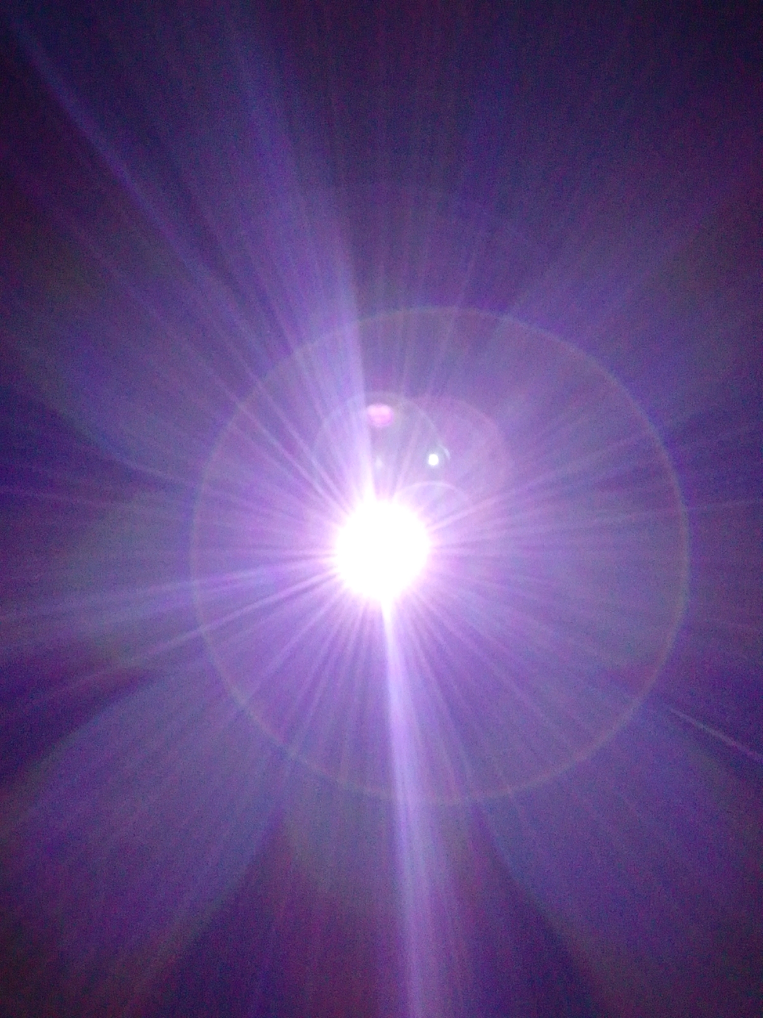 The image shows a bright light source with visible rays emanating outwards, creating a starburst or lens flare effect. The dominant color is purple, with variations in intensity and hue due to the light. The background is a dark, possibly night sky, with some subtle gradations of color. There are also some circular or oval light reflections, indicating the light source is likely artificial, such as a powerful lamp or spotlight, or a very bright celestial object like the sun or moon captured with a lens flare. The image lacks any discernible details of people, objects, actions, interactions, or a specific location within Serti, Nigeria. The abstract nature of the image makes it impossible to provide a forensic description related to a specific scene.