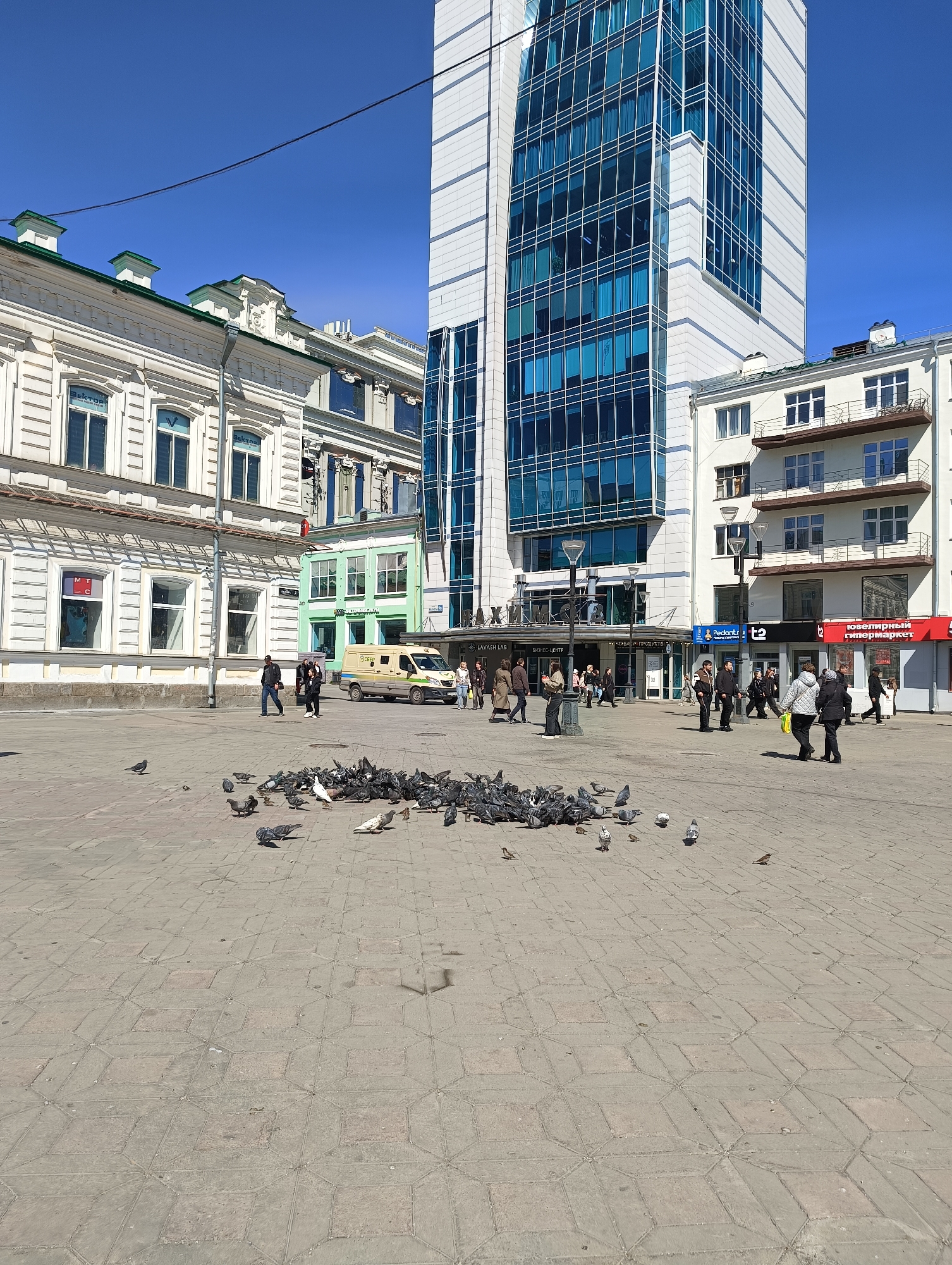 A square in Yekaterinburg, Russia, is paved with patterned concrete tiles, upon which a large group of pigeons gathers. People walk across the square in various directions. A yellow armored van is parked in the background. Buildings surround the square, including a modern glass-fronted tower and older, ornately decorated buildings. Signs in Russian are visible on some buildings. The sky is clear and blue.