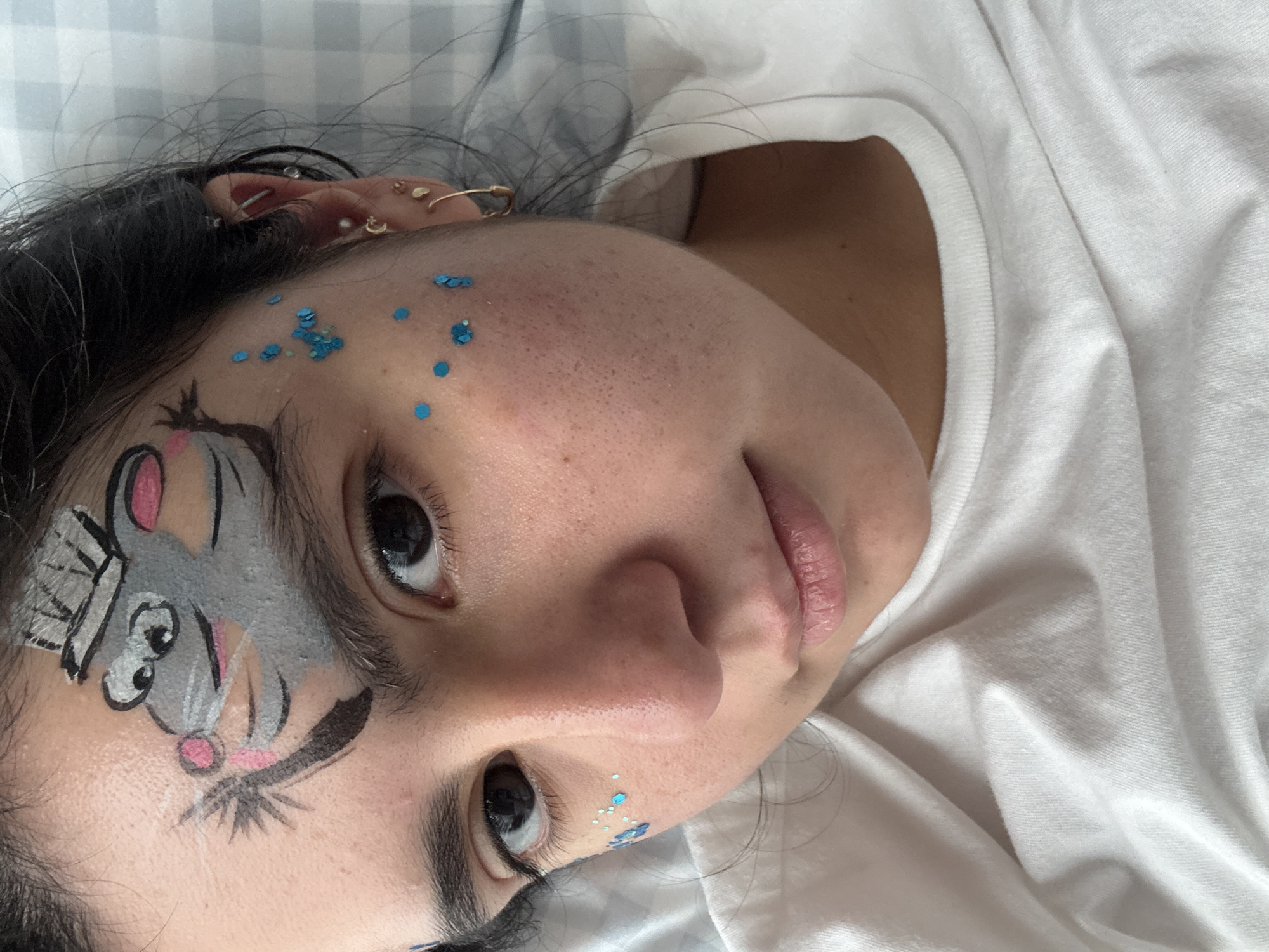 A young woman with dark hair lies on her back, wearing a white t-shirt. Her face is adorned with face paint and glitter. The face paint depicts a cartoon mouse wearing a hat, positioned on her forehead above her left eye. Blue glitter is scattered across her cheeks and temples. She has dark eyes and appears to be looking directly at the camera. The background is a white and gray checkered pattern, possibly a sheet or blanket. The scene appears to be indoors.
