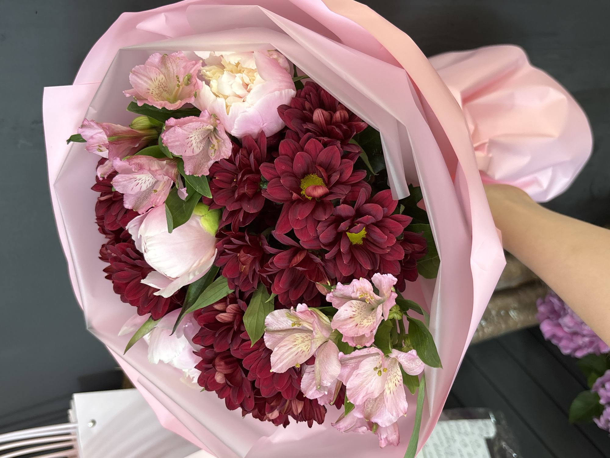 A close-up, eye-level shot shows a hand holding a bouquet of flowers wrapped in pink paper. The bouquet features deep red chrysanthemums, pale pink alstroemeria, and light pink peonies. The background is dark and blurred, with a hint of a white object on the left. The scene is indoors, possibly in a flower shop in Chaikovsky, Russia, given the stylistic choices often seen in floral arrangements from that region.