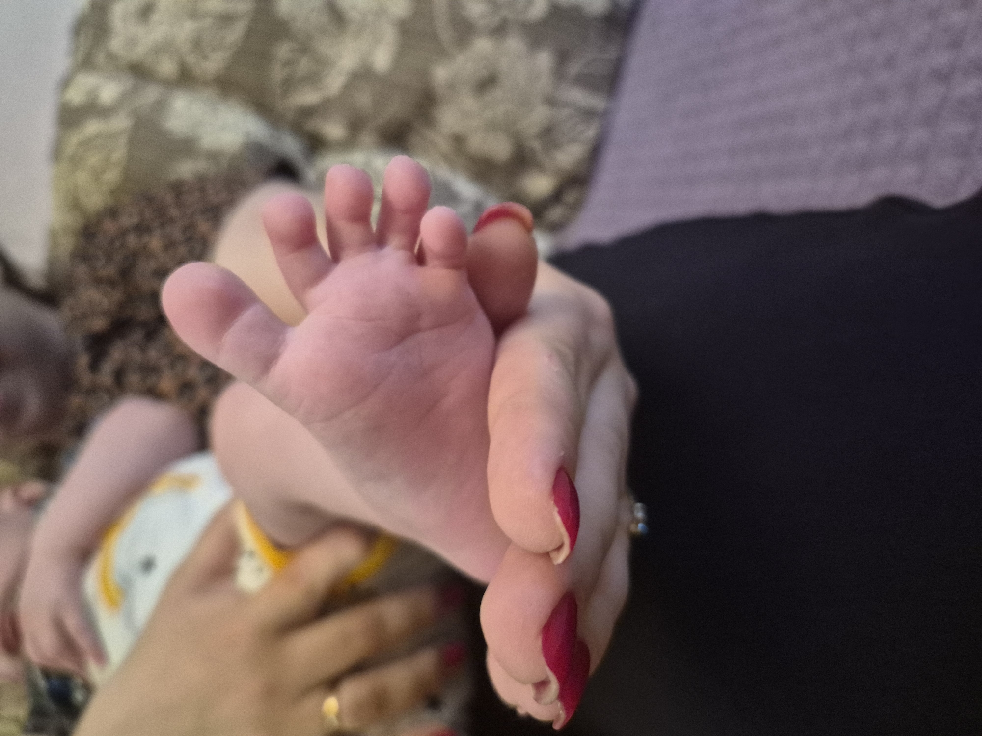 The image shows an infant's foot held by a woman's hand. The woman's fingernails are painted a dark red color. The infant is wearing a diaper with yellow trim and a print of cartoon characters. The infant is lying on a surface with a floral pattern. The background is blurred, but a dark purple surface is visible. There is no indication of the location being Tobolsk, Russia.