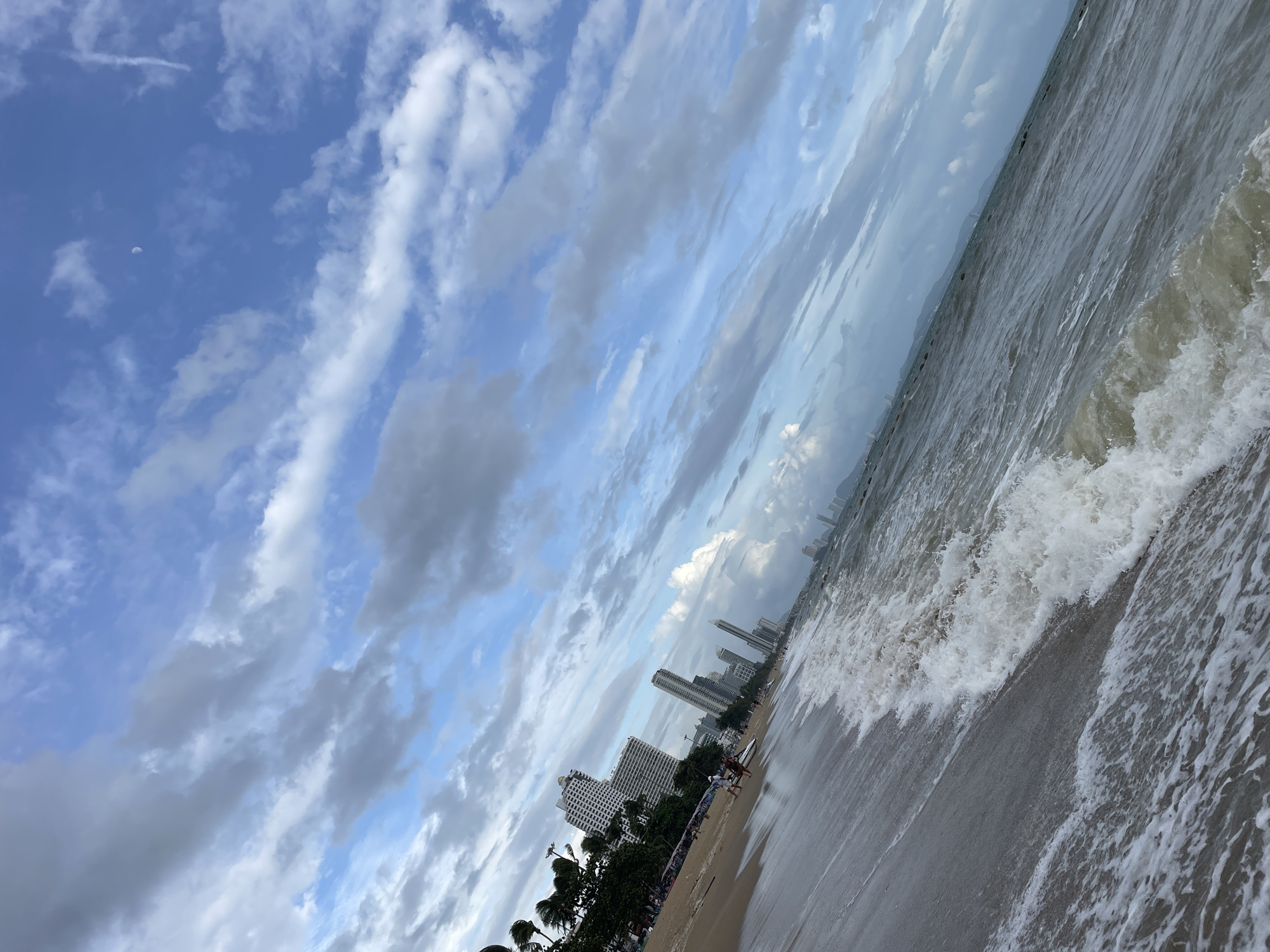 A tilted, eye-level, wide shot captures a dynamic view of Pattaya City, Thailand, from the sandy shoreline. The foreground is dominated by the ocean, with frothy waves rolling onto the wet sand. The water is a murky grey-brown, suggesting depth or sediment. In the midground, the sandy beach is visible, lined with palm trees and several tall, white high-rise buildings that form the city's skyline. A few individuals are present on the beach, seemingly engaged in leisure activities. The background is a vast expanse of blue sky, adorned with dramatic, greyish-white clouds that stretch diagonally across the frame, adding a sense of movement and depth to the scene. A crescent moon is faintly visible in the upper right portion of the sky. The overall impression is of a bustling urban coastline under a cloudy sky.