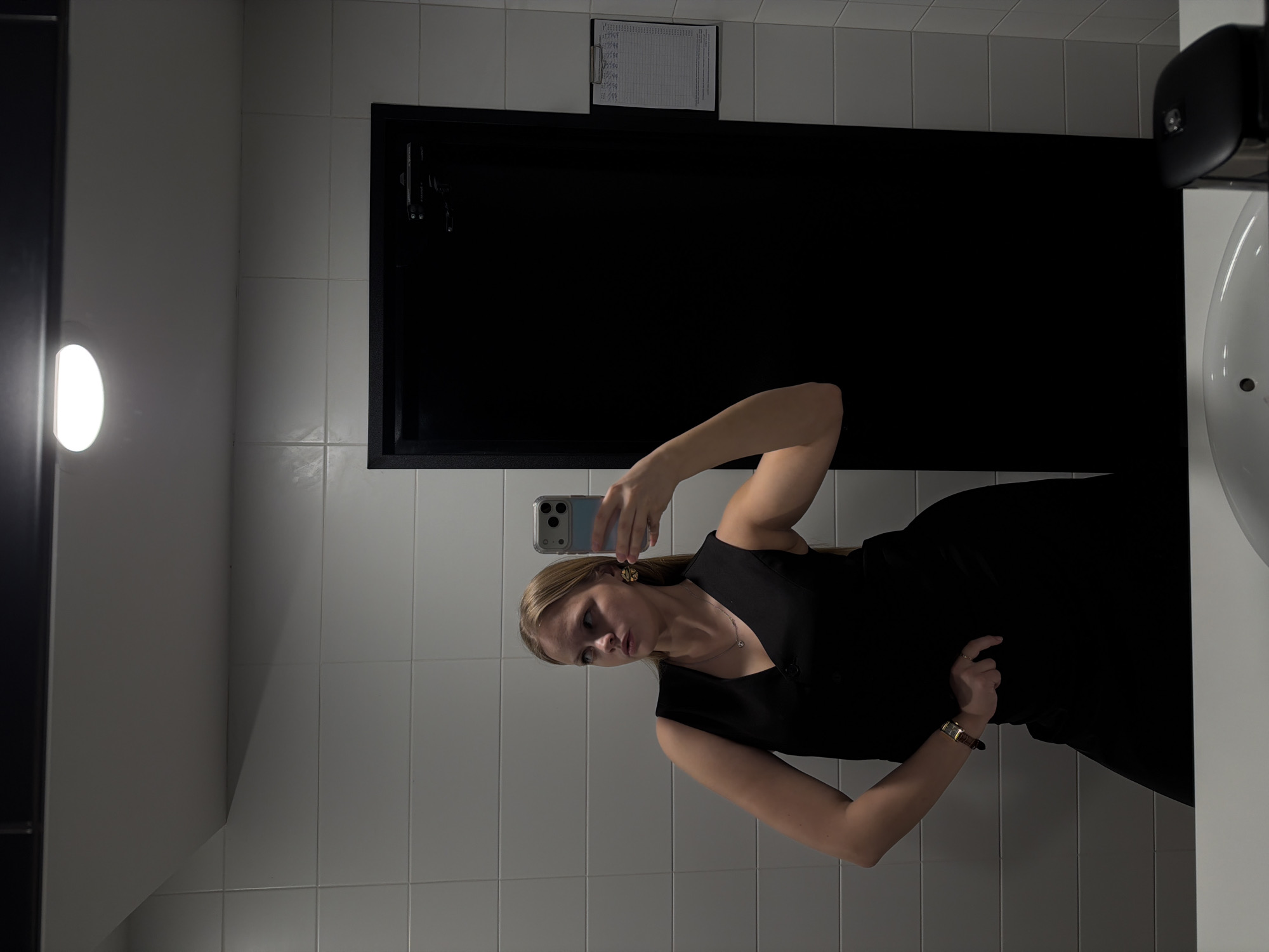 A woman is taking a mirror selfie in a bathroom. The bathroom has white tiled walls, a black door, and a sink. A schedule or document is attached to the wall above the door. The location of this image is not in Izhevsk, Russia.