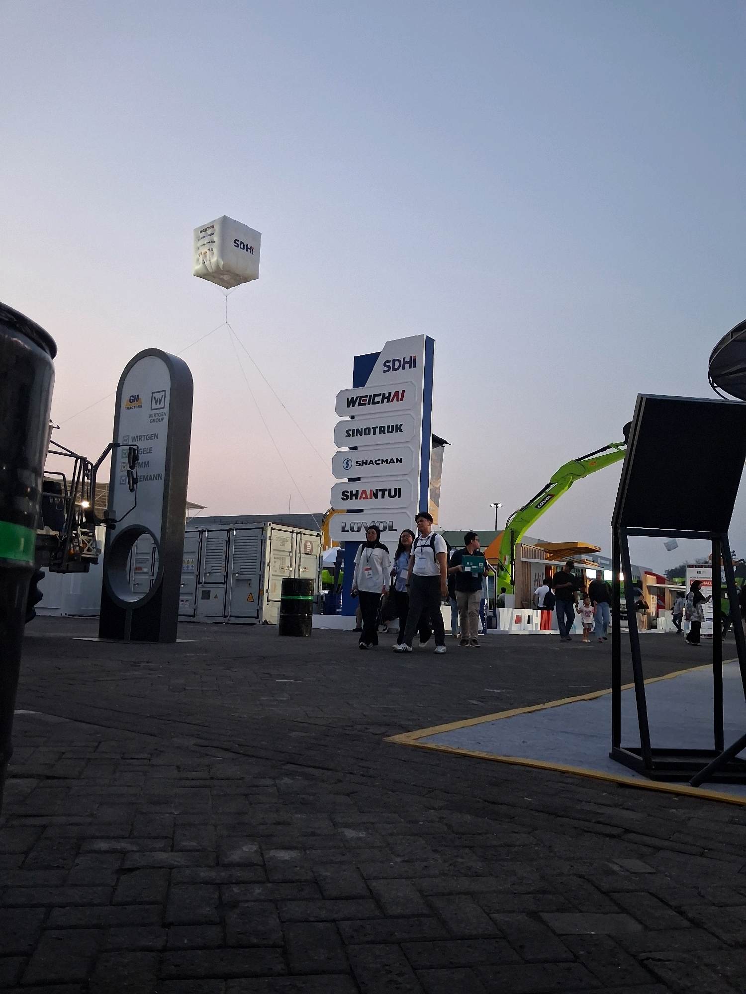 This is an eye-level, full-shot photograph of an outdoor exhibition or event. In the foreground, a dark, paved ground is visible, composed of rectangular stones. To the left, a portion of a large, black, cylindrical object with a green stripe is in the frame. Behind this, a tall, dark grey, oval-shaped structure with a circular opening is prominently displayed. It features a white surface with branding including "GM TRACTORS" and "WIRTGEN GROUP" along with a list of names. A white cube-shaped balloon with text like "SDHI" and "WEICHAI" floats in the sky above.
Further back, a large white signpost with multiple brand names such as "SDHI," "WEICHAI," "SINOTRUK," "SHACMAN," "SHANTUI," and "LOVOL" is erected. Several people are walking in the middle ground. A bright green excavator is parked behind the signpost, with its arm extended. Shipping containers are visible behind the oval structure and the signpost. The background shows a dimly lit sky, indicating either early morning or late evening. The scene appears to be set in an open area, possibly a fairground or exhibition center, in Jakarta, Indonesia.