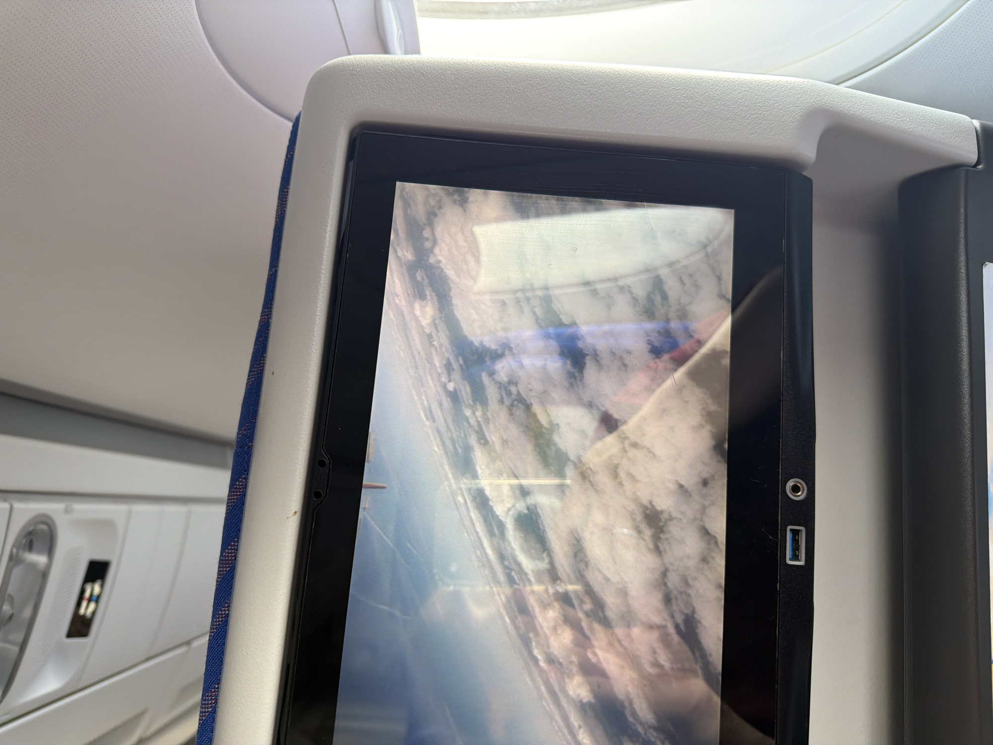 The image captures an interior view of an airplane cabin, focusing on an in-flight entertainment (IFE) screen attached to the back of a seat. The screen displays an aerial view, oriented sideways, showing a landscape of clouds, land, and what appears to be a body of water or coastline. Reflections are visible on the screen's surface, possibly from ambient cabin lighting or a window.
On the right bezel of the IFE screen, a circular headphone jack and a rectangular USB-A port are clearly visible. The screen is housed within a light gray plastic frame, which is part of the seat-back structure. A blue fabric strip with a subtle reddish pattern runs along the left side of the screen's frame, providing a visual accent.
To the left of the screen, parts of the airplane's interior ceiling are visible, including a light-colored panel with a dotted texture and a portion of an overhead compartment or light fixture with a reflective handle. Another seatback or partition is partially visible on the far right.
The scene depicts a passenger's experience during air travel, likely viewing content on the IFE system. No people are visible in the frame. The view on the screen suggests it is daytime. No discernible text is visible on the screen or surrounding cabin elements. The contextual location provided for the image is Parshino, Russia.