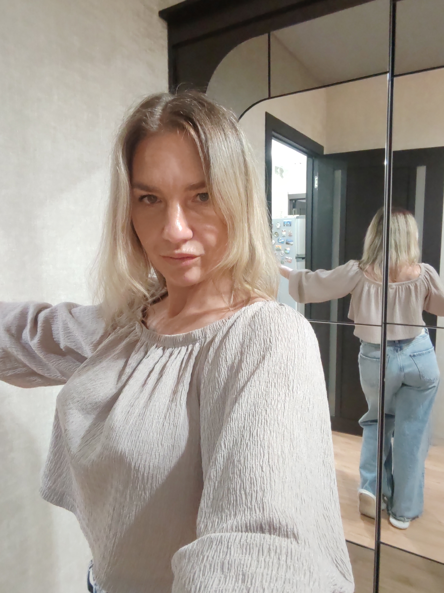 A fair-skinned woman with blonde hair stands in what appears to be a residential interior, likely a bedroom or hallway. She wears a textured, light-colored, long-sleeved shirt and light blue jeans. She is facing the camera, while her reflection in a mirrored closet door reveals her back. The reflection shows her standing with her arms extended to the sides.
Behind her, visible through the reflection in the mirror, is a refrigerator decorated with magnets. The location is possibly in Petropavlovsk-Kamchatsky, Russia, given that the user instructed to describe the image as if it were taken there. However, no specific details in the image confirm this location.
