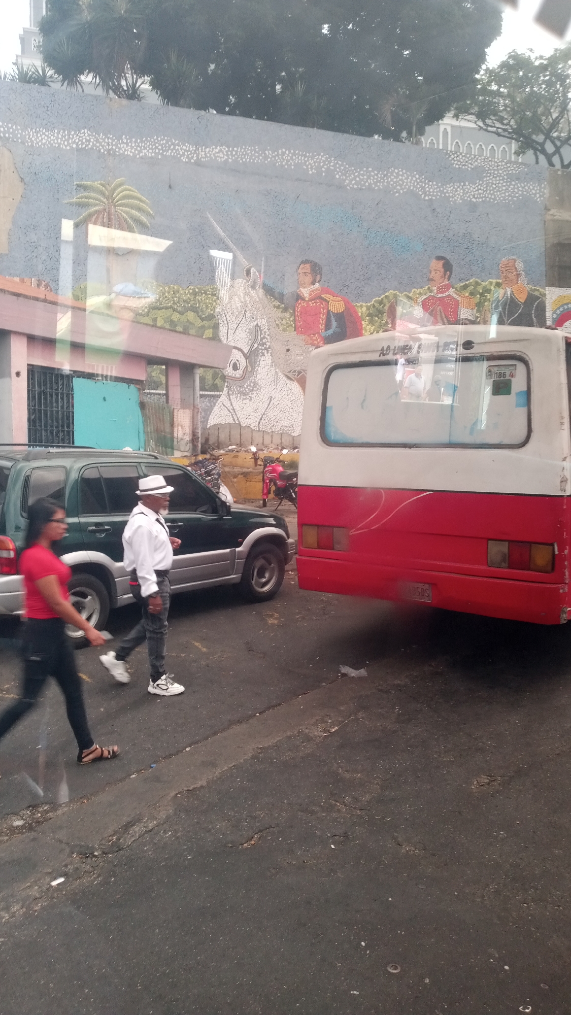 The photograph captures a street scene in an unidentified urban location, possibly in Unknown, Unknown. The foreground shows a paved road with visible wear and tear. A woman in a red shirt and black pants is seen walking to the left, and a man wearing a white hat, shirt, and gray pants is walking to the right. To the left, a green and silver SUV is parked. A red and white bus obscures part of the background. Behind the vehicles and pedestrians, there's a wall adorned with a mural depicting historical figures and a white horse. A palm tree is also present in the mural. The background includes trees and buildings.
