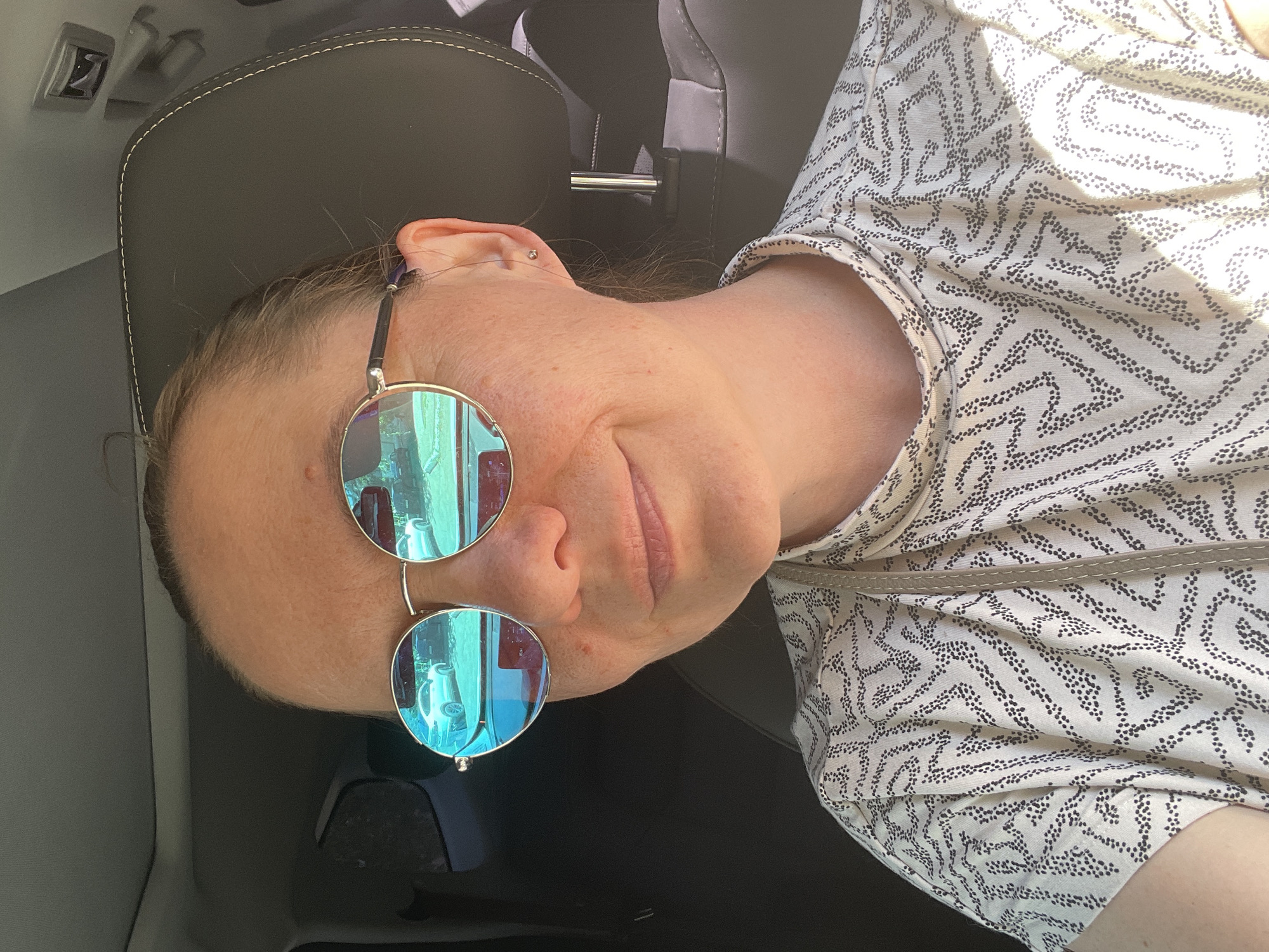 A woman is sitting in the driver's seat of a car, wearing round, mirrored sunglasses with blue lenses, a white and black patterned t-shirt, and a simple stud earring. The sunglasses reflect images of cars and buildings. The interior of the car is visible, including the headrest and a dark dashboard. The lighting suggests it is daytime and sunny. The background is out of focus.
