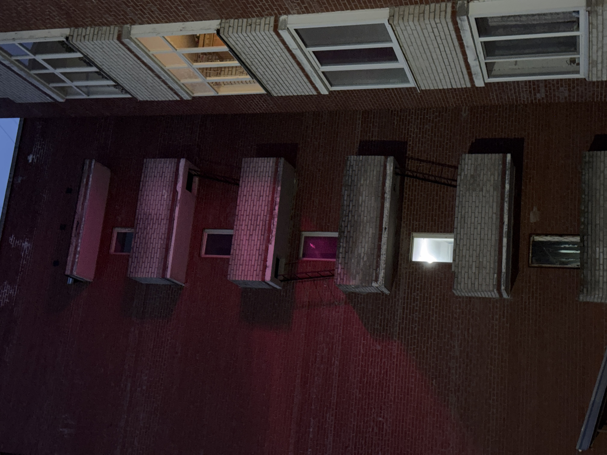 A photograph of a building facade at night shows a brick wall with several windows and architectural protrusions. The building's brickwork is a deep red hue, illuminated by varying light sources. A row of windows is visible at the top, showing rectangular panes with white frames. Below the windows, there are architectural elements that jut out from the wall, constructed of bricks and illuminated by different lights from within the building, creating a contrast of shadows and light. The overall impression is of an urban setting with a structured architectural style.