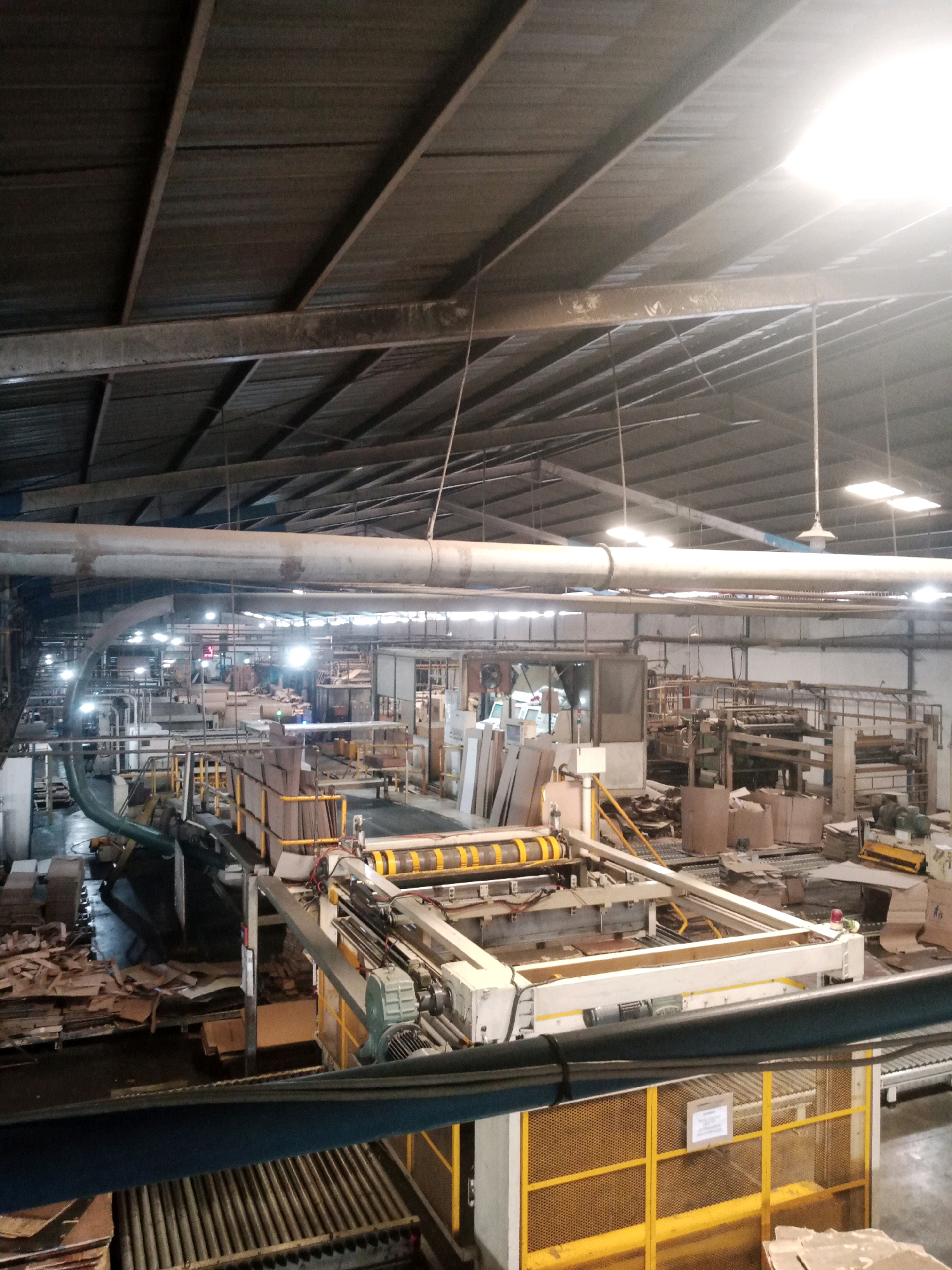 The image shows an interior view of a factory in Balaraja, Indonesia, likely involved in the production or processing of cardboard materials. The scene is industrial, with a high ceiling supported by metal beams and corrugated metal roofing. Large ventilation pipes and lighting fixtures are suspended from the ceiling.
In the foreground, a machine with yellow and white components is visible, featuring rollers and metal framing. This machine appears to be part of a conveyor system or production line. Various stacks of cardboard and raw materials are scattered throughout the factory floor. In the midground and background, more machinery, shelving units, and cardboard boxes are present, indicating a busy operational environment. The lighting is uneven, with some areas brightly illuminated by overhead lights and others in shadow. No people are clearly visible in the image. The overall impression is one of a functional, albeit somewhat cluttered, manufacturing facility.
