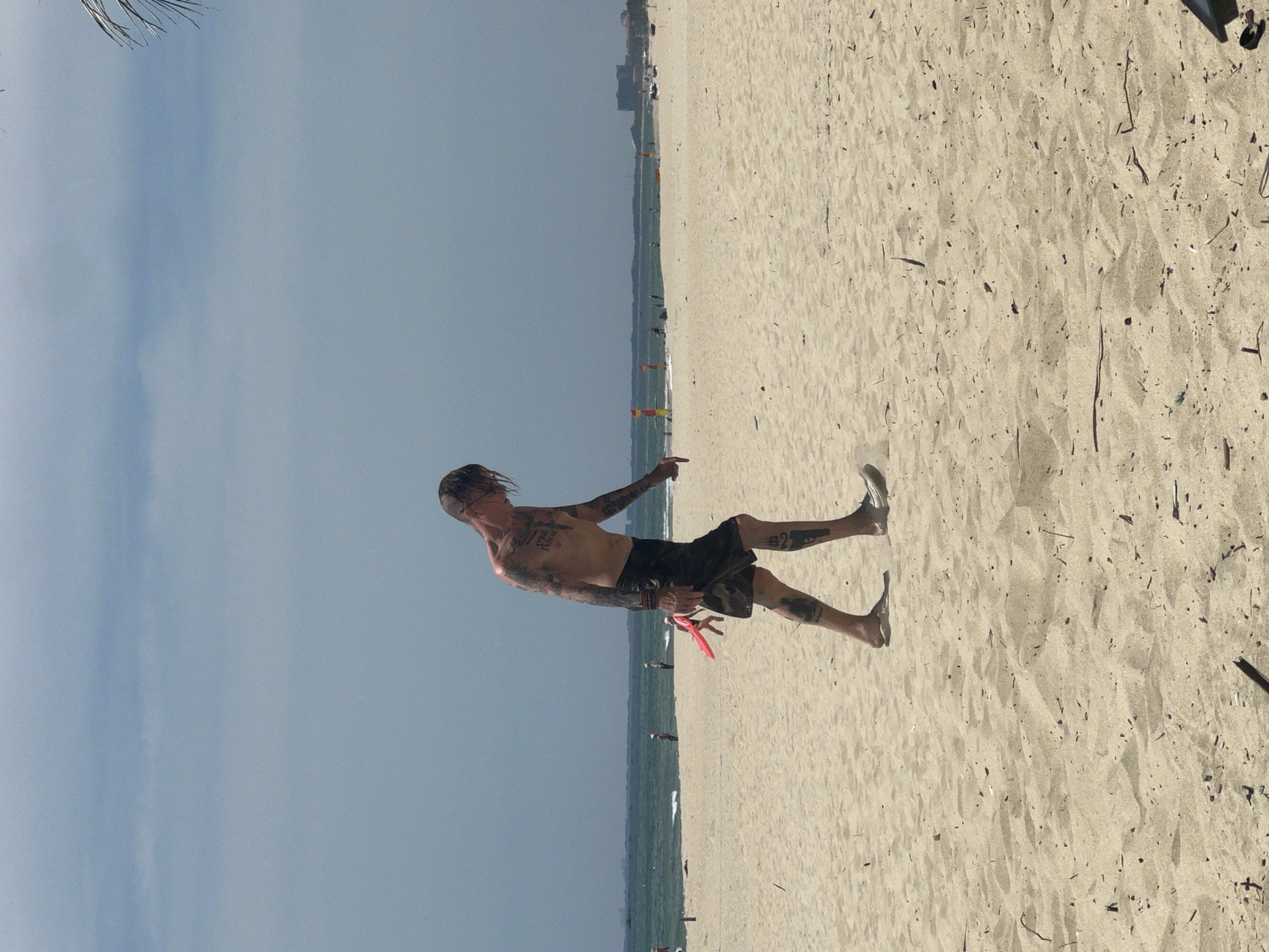 A man with extensive tattoos on his torso and arms walks along a sandy beach. He is wearing dark patterned shorts and appears to be barefoot on the left foot and wearing a light-colored shoe on the right foot. In his right hand, he is holding a small, bright red object. In the background, the ocean meets a hazy horizon, with a few buildings visible in the distance, suggesting an urban coastal setting in Đà Nẵng, Vietnam. The sky is overcast with a pale blue hue. Several other indistinct figures are scattered on the beach and in the water in the distance. The sand is light brown and shows some scattered debris.