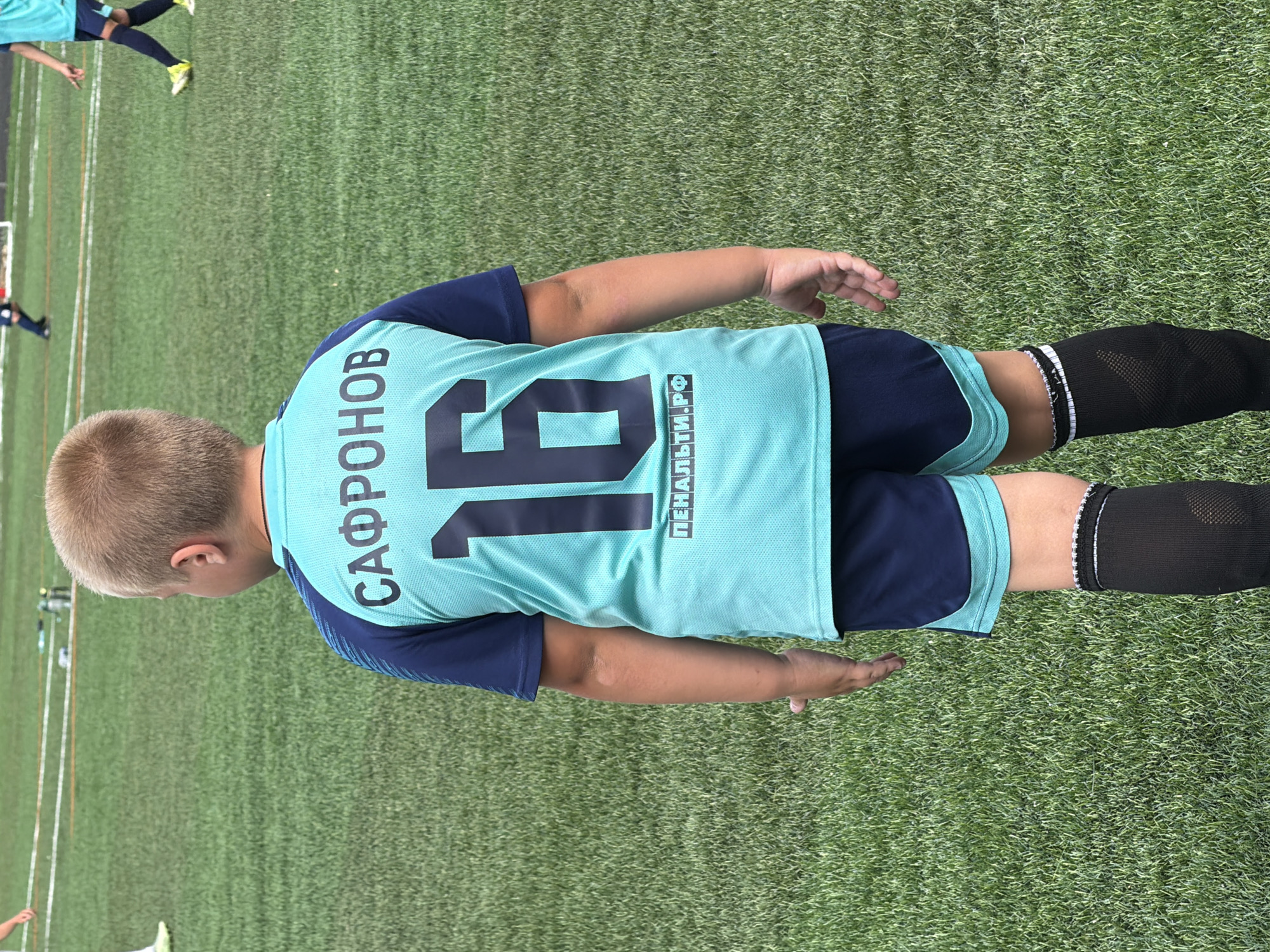 A young boy, seen from behind, stands on a green grass soccer field. He wears a light blue and navy blue soccer uniform, consisting of a short-sleeved jersey with the name "САФРОНОВ" and the number "16" printed in dark blue, and navy blue shorts with light blue trim. He also wears black knee-high socks. The setting is a sports field, with white lines marking boundaries and other players and equipment visible in the background. The image appears to have been taken outdoors in Bykovo, Russia, given the context provided.