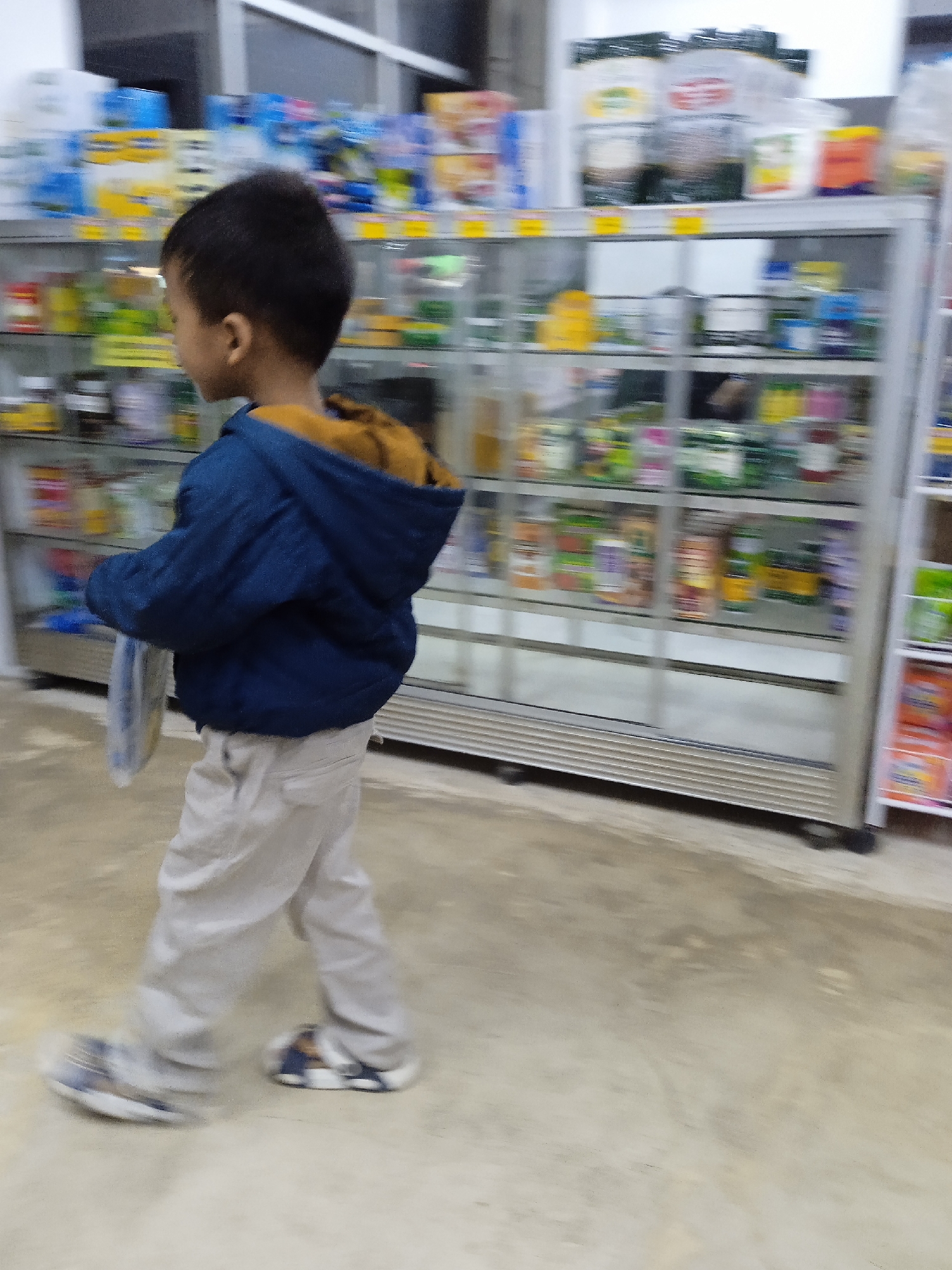The image depicts a young boy walking through a small shop or convenience store in Kebonsari, Indonesia. The boy is seen from a slightly elevated side-back perspective, moving from left to right across the frame. He wears a dark blue hooded jacket, which appears to have a brown or gold inner lining, light-colored or beige pants, and sneakers. Significant motion blur is visible on his feet, indicating movement. In his left hand, he holds a light-colored, somewhat translucent object, likely a plastic bag or an item wrapped in plastic. His head is turned slightly, showing the back and side of his head with dark hair.

The background features multiple glass-fronted display cases or shelves, characteristic of a retail setting. These shelves are stocked with various packaged goods, though specific items are indistinct due to motion blur and distance. They appear to be an assortment of food products, beverages, or household items. The lighting within the store is moderate, suggesting an indoor environment. The floor is a light-colored, possibly concrete or tiled surface, showing some minor scuffs. There is no clearly legible text visible on any products or signage within the image. The scene captures a momentary action, with the blur conveying the boy's movement.