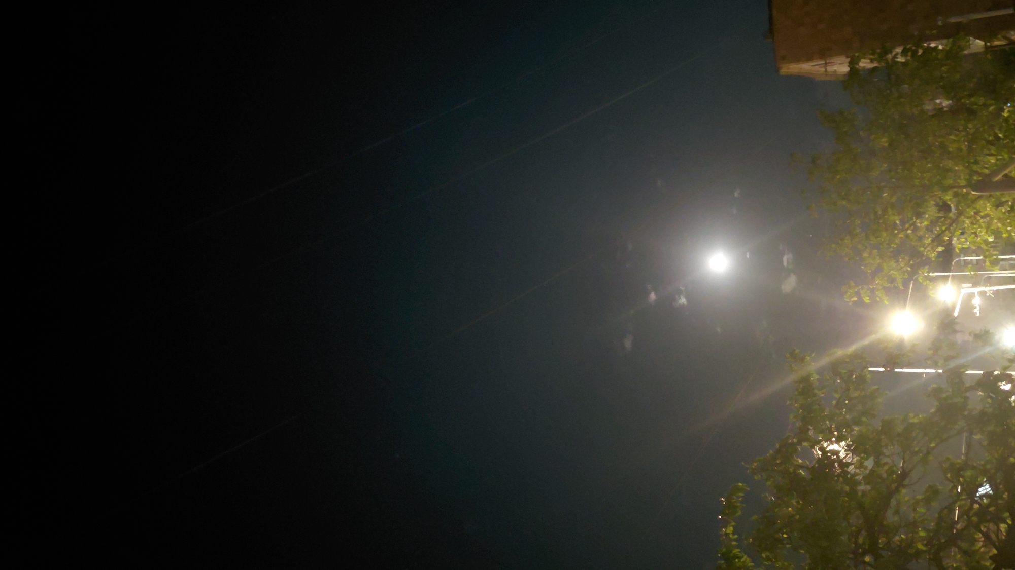 The image depicts a night scene in the city of Abovyan, Armenia, featuring a moonlit sky above trees and streetlights. The moon, a bright, luminous orb, is visible in the upper center of the frame, partially obscured by wispy clouds. Below the moon, several streetlights cast a warm, yellow glow, illuminating the surrounding trees and foliage. The trees, with their dense green leaves, occupy the lower portion of the image, framing the moonlit sky. A building, likely an apartment block, is partially visible on the right side of the frame, its facade lit by the streetlights. The overall atmosphere is tranquil and serene, with the natural light of the moon and the artificial illumination of the streetlights creating a soft, diffused light. The sky is a deep, inky blue, with faint streaks of light, possibly from distant stars or reflections. The image appears to be taken from a street-level perspective, offering a view of the urban landscape under the night sky.