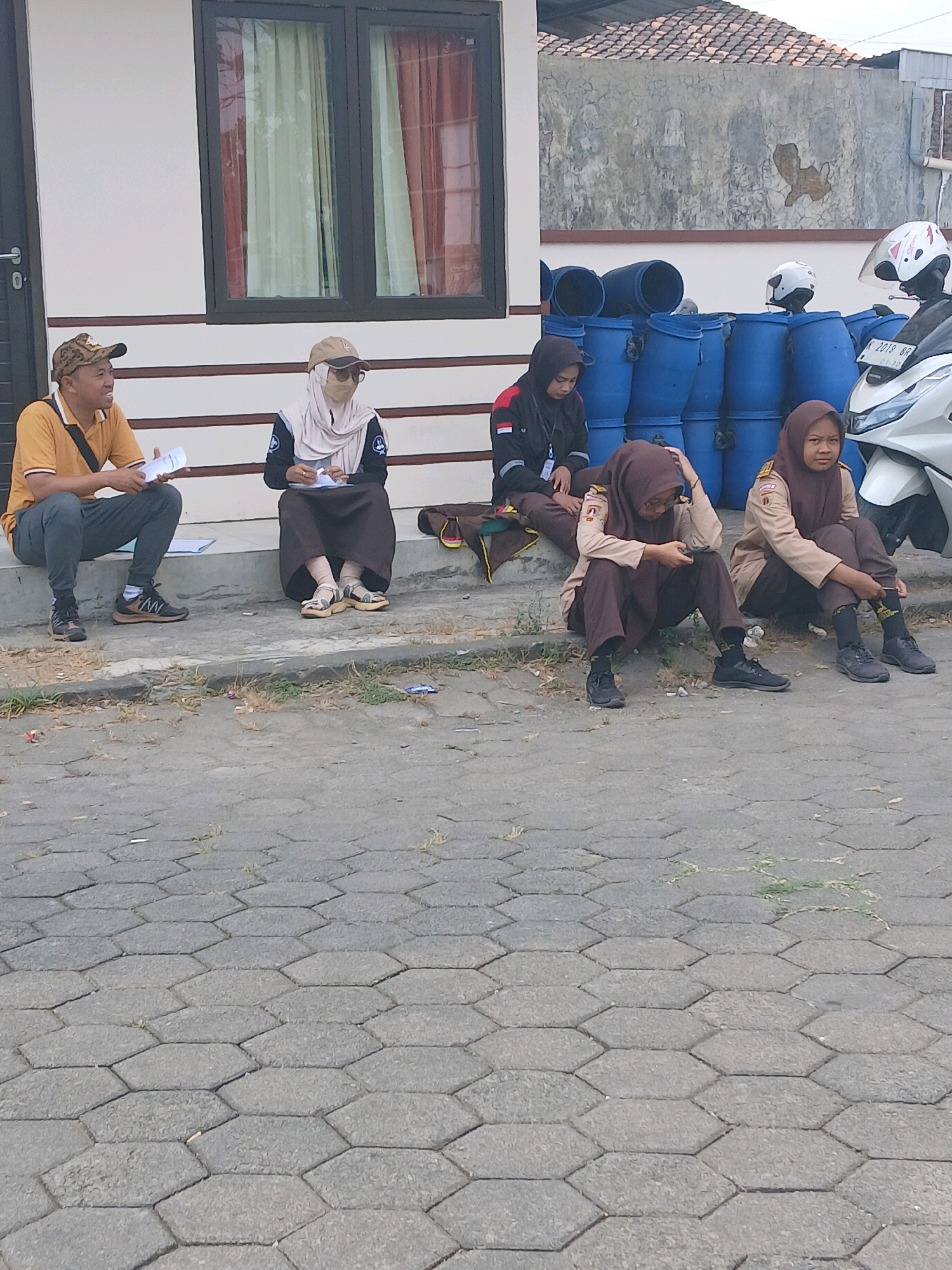 Six individuals, five appearing to be students in school uniforms and one adult male, are seated outdoors on a paved area in front of a building. The adult male, wearing a yellow shirt and a patterned cap, is sitting on the steps of the building and holding papers. Next to him, on the steps, a student in a light brown hijab and a dark top is also holding papers. Further to the right, on the ground, two other students in similar uniforms are seated, one looking down at their phone. Another student, also in uniform, is seated to the left, wearing a black jacket with red accents. A fifth student in uniform is seated on the ground to the far right. In the background, large blue barrels are stacked, and a white motorcycle with a helmet is visible. The ground is paved with hexagonal grey stones, and sparse vegetation grows between them. The building behind them has white walls and dark-framed windows. The scene suggests a waiting or resting period for the group. The location is identified as Jepangpakis, Indonesia.