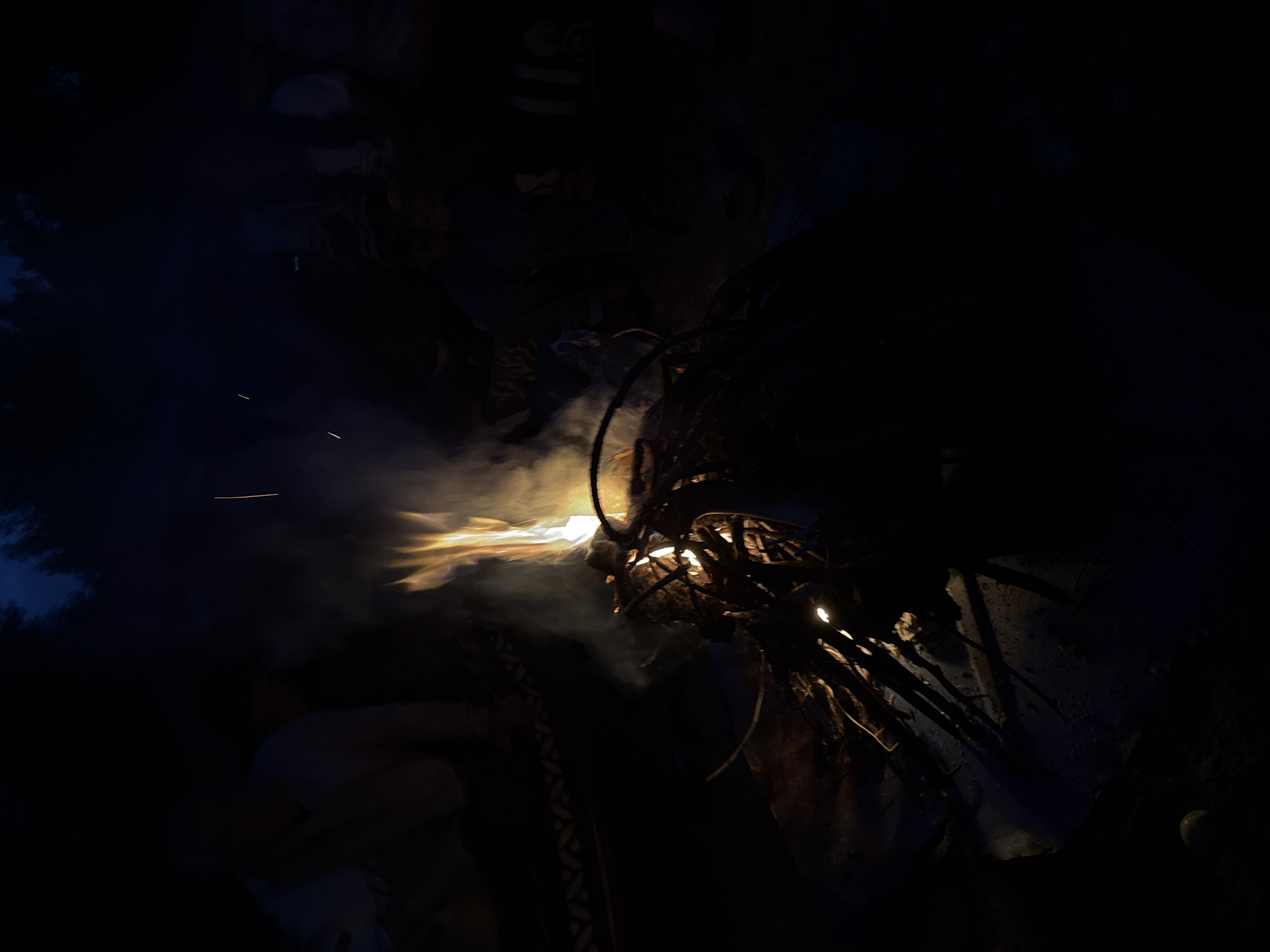 The image depicts a nighttime scene in Sevan, Armenia, centered around a campfire. The fire is burning brightly with flames reaching upwards and emitting smoke. Around the fire, there are several people partially visible, silhouetted against the darkness and illuminated by the firelight. They appear to be gathered around, possibly for warmth or conversation. The environment is dark, with the sky above appearing deep blue or black, suggesting it is late at night or early in the morning. The ground around the campfire is covered with what appears to be dirt and debris. The overall atmosphere is one of a relaxed, outdoor gathering.