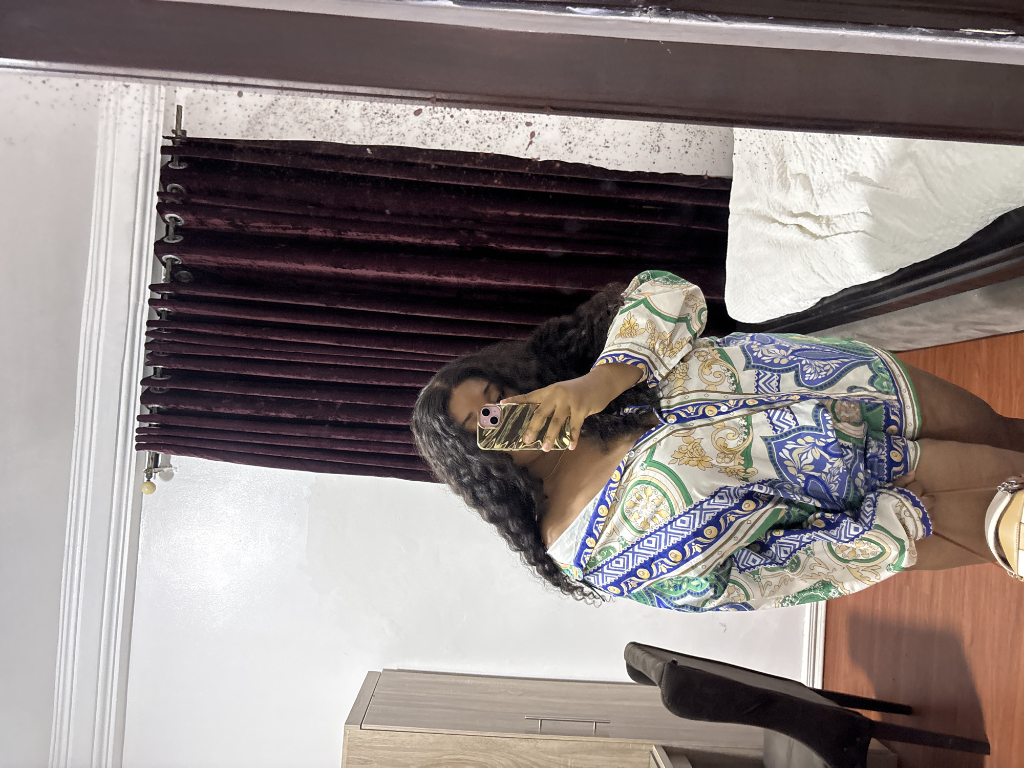 A woman is taking a selfie in a room, using a gold-colored phone. She is wearing a blue, white, green, and gold patterned top. The background includes a burgundy curtain, a white wall, a chest of drawers, and a chair. The reflection suggests the photo is taken in a mirror. The location is identified as Port Harcourt, Nigeria, based on contextual information.