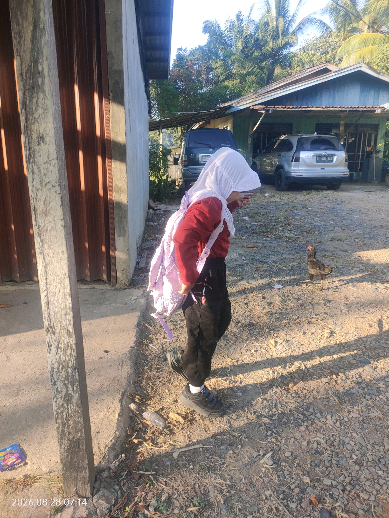 A person wearing a white hijab and a red jacket walks on a gravel path in front of a building. The person is carrying a light purple bag over their shoulder. A chicken stands on the ground to the right of the person. Behind the person and chicken is a light blue house with a corrugated metal roof, and two parked cars. The ground is covered with gravel, dirt, and dry vegetation. The image is dated 2026.08.28 07:14. This scene is likely in Kuaro, Indonesia.