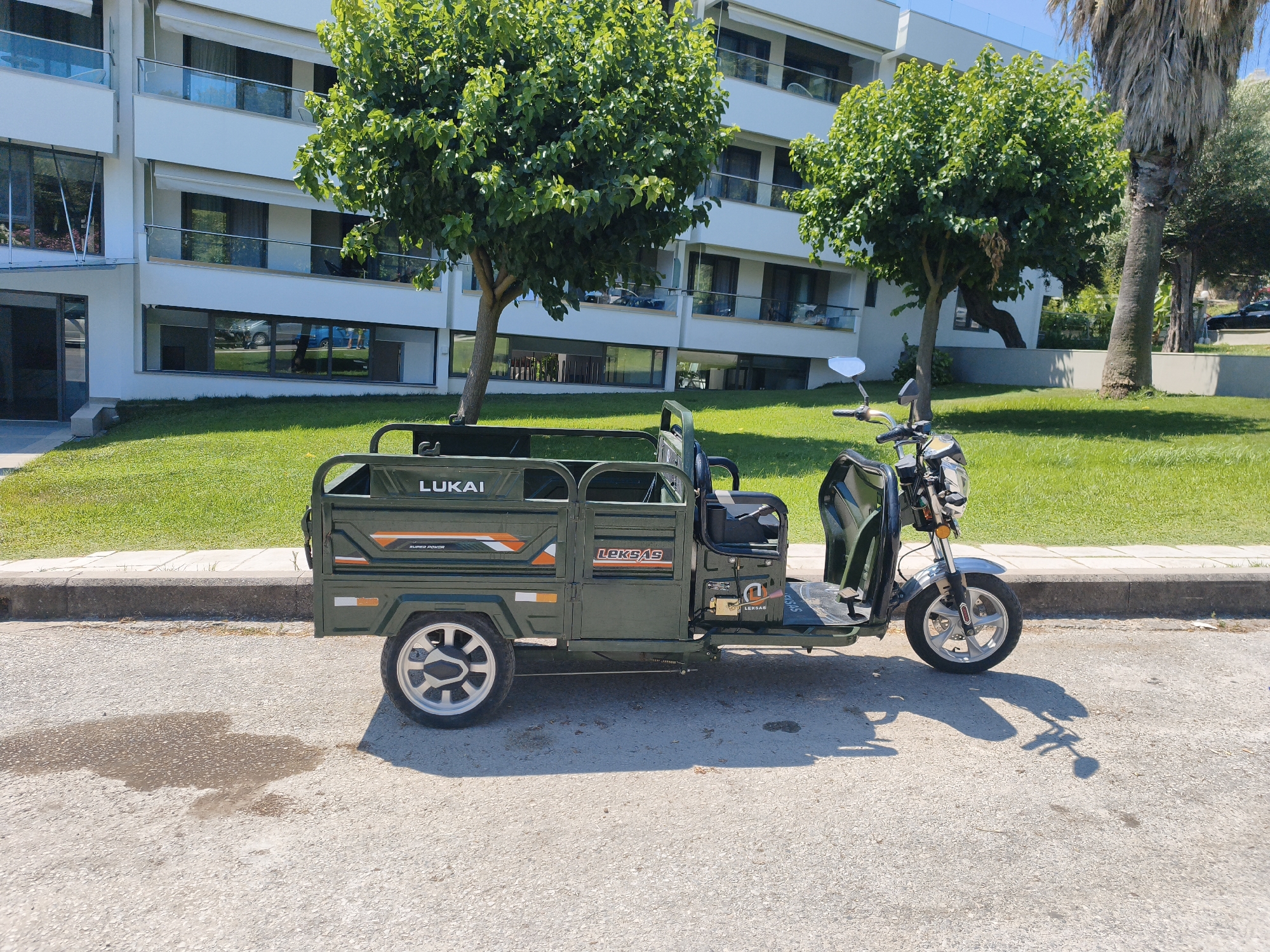 A three-wheeled green cargo scooter, identified by the "LUKAI" and "LEKSAS" branding on its side, is parked on a paved surface. The scooter has a driver's seat, a cargo bed in the rear, and a metallic front fender. It is positioned in front of a grassy area and a modern white building with balconies and tinted windows. Two trees with green foliage are visible in the foreground, one on the left and one behind the scooter. The environment appears to be a sunny day, casting shadows on the pavement. The location is the Municipal Unit of Kassandra, Greece. There are no people visible in the image.