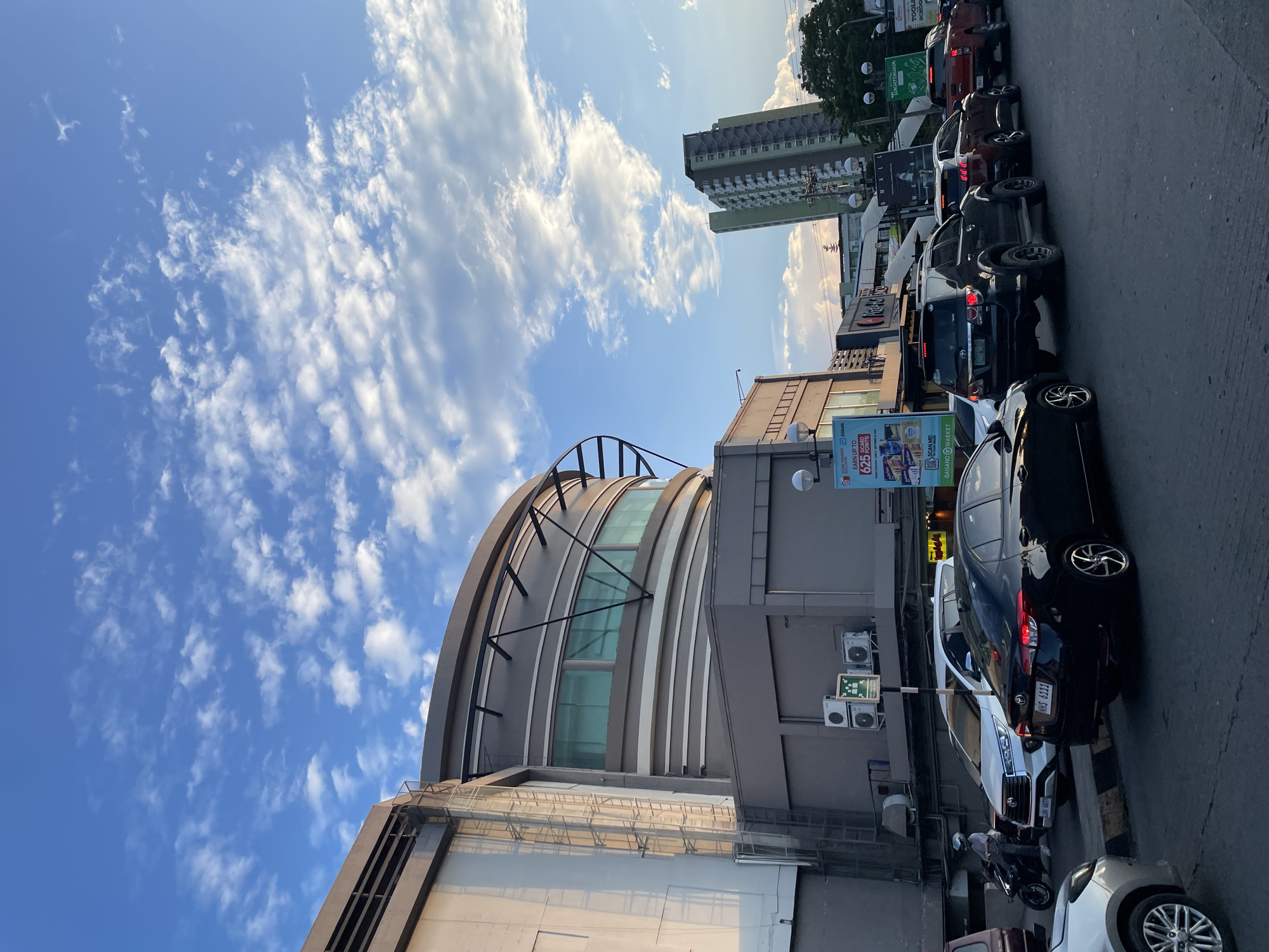 The image depicts a traffic scene in front of a large, modern building with a curved facade, likely a mall, in Davao City, Philippines. The sky is partly cloudy with a blend of blue and white, suggesting it might be late afternoon or early evening. Several vehicles, including cars, SUVs, and a motorcycle, are lined up on the road, indicating a traffic jam. The building features large windows and visible air conditioning units. Billboards advertising various products and services are also present. In the background, a tall, green-tinted building stands out against the sky. The overall scene is characteristic of a bustling urban environment in the Philippines.