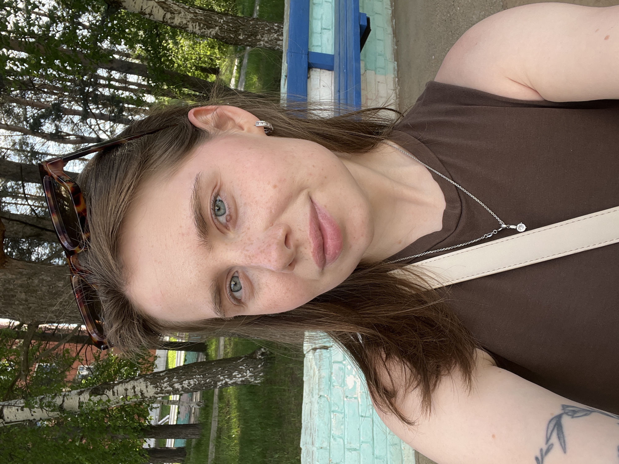 A woman with fair skin and brown hair is centered in the frame, wearing a brown sleeveless top and a silver necklace with a small pendant. Sunglasses are perched on her head. The location is Chaikovsky, Russia. In the background, birch trees, green grass, and a brick structure are visible. A bench painted with blue and turquoise stripes is partially visible in the background. The image appears to be a selfie.