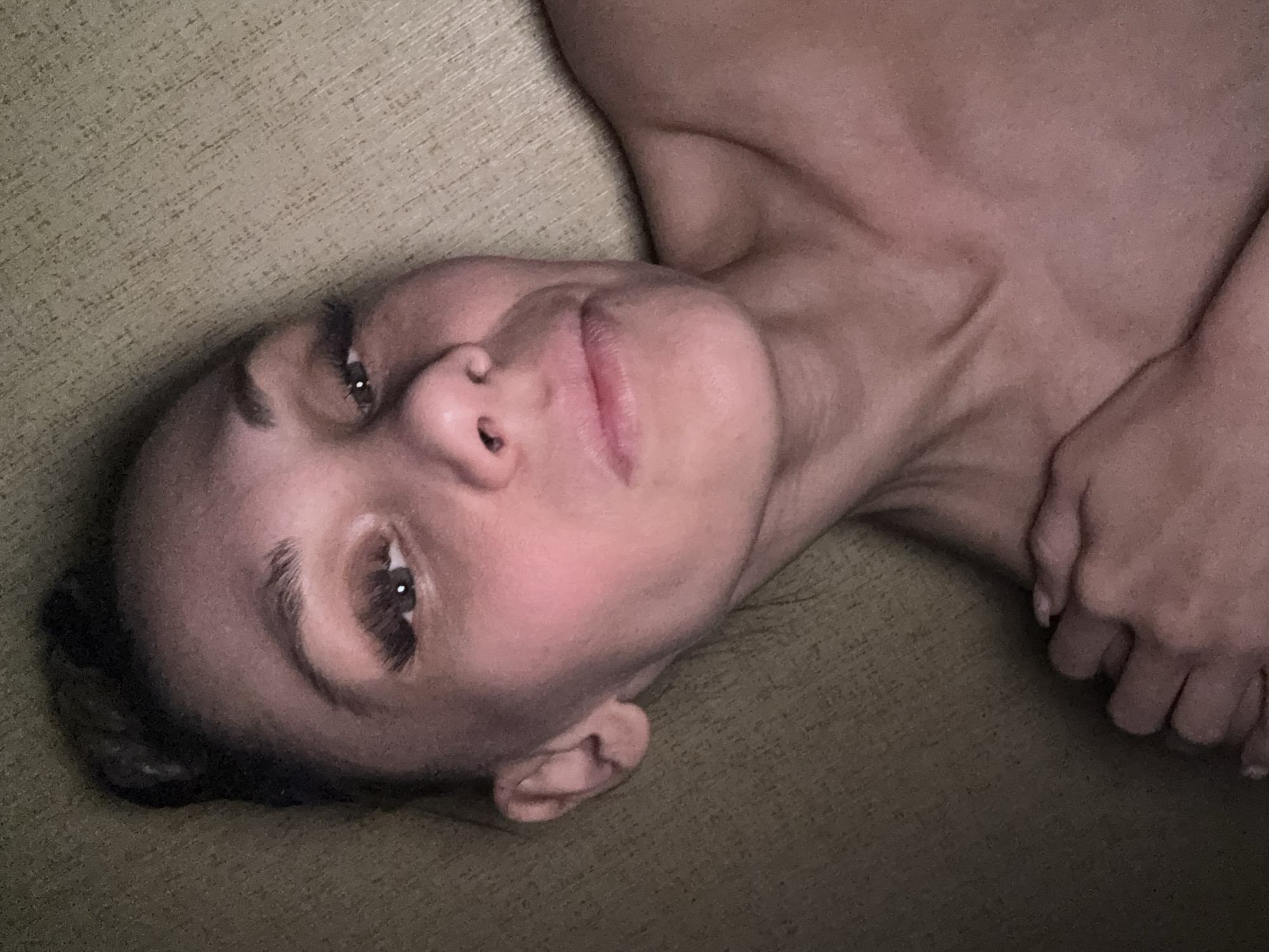 A woman is lying down on a tan, textured surface. The image is taken from an overhead angle, with the woman's head tilted to the left. Her face is visible, displaying her left eye and the bridge of her nose, with light skin with a pinkish hue. Her hair is dark and appears to be pulled back. Her neck and shoulder are also visible, and she is holding her hands in front of her. The location is a plain, indoor setting, devoid of distinguishing landmarks and city-specific elements to establish a precise location. Therefore, stating the location is Unknown, Russia, is speculative without further evidence.