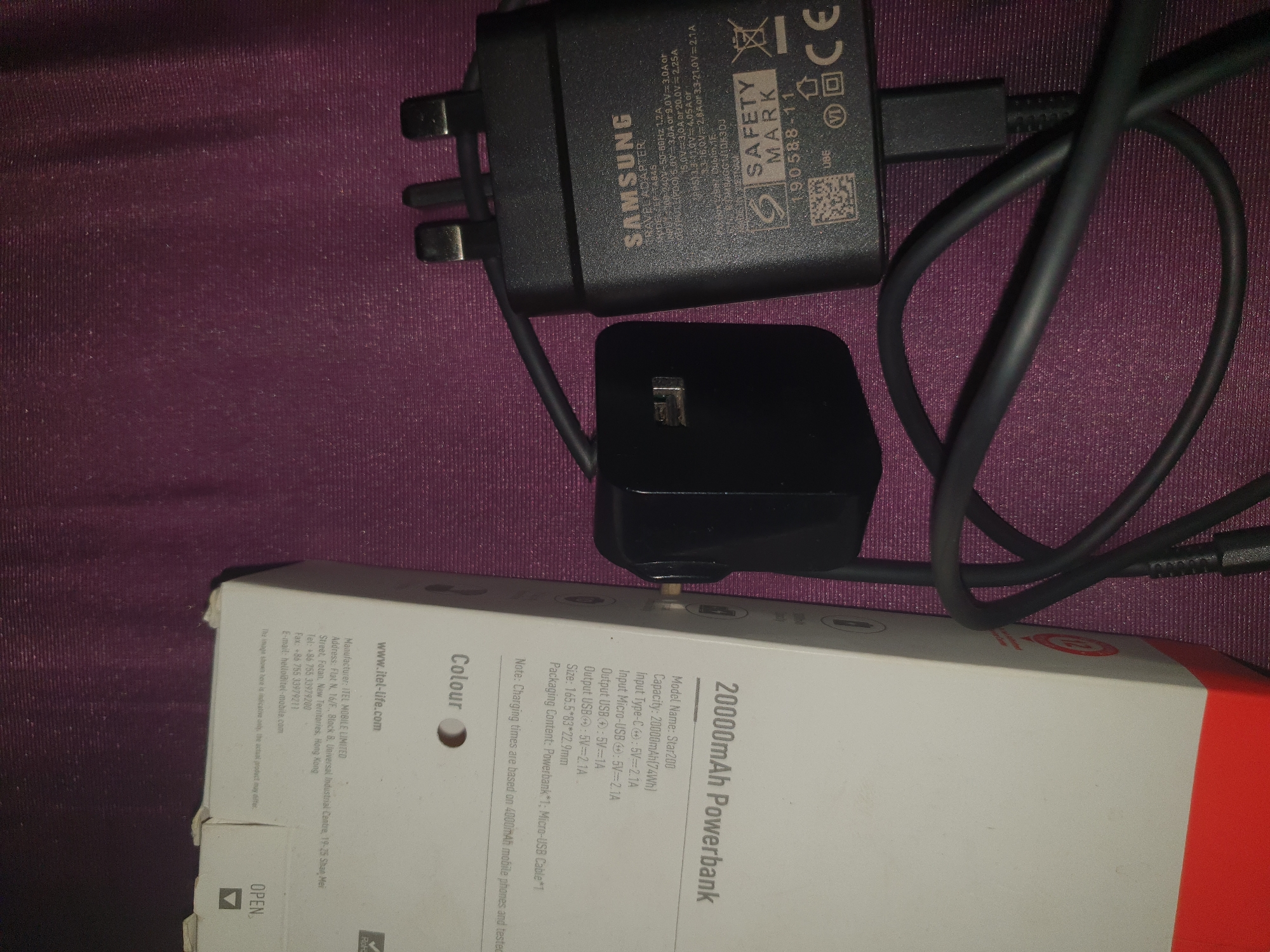 The image shows a black Samsung travel adapter and a white box with "20000mAh Powerbank" printed on it, along with a black USB cable connecting the two. The box also has "Colour" and "Model Name: Star200" printed on it. The background is a dark purple textured surface. The Samsung adapter has text indicating it's for travel and lists output specifications. The scene is indoors with artificial lighting. There are no people or specific environmental features to identify a city or location.