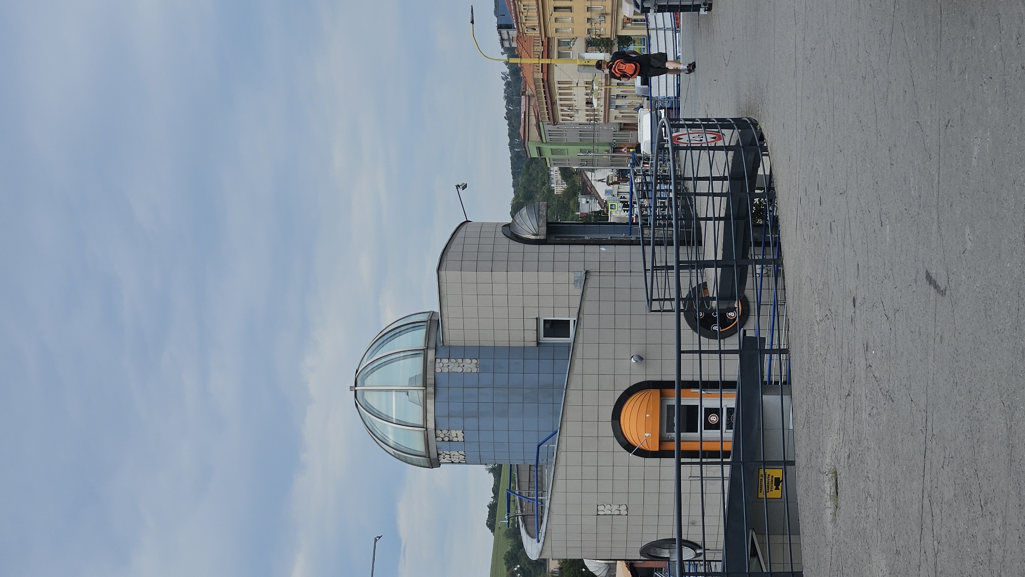 The image depicts the exterior of a modern building with a distinctive domed roof, located in Prešov, Slovakia. The building features a curved facade covered in grey tiles, with accents of blue tiles and circular decorative elements. An orange-arched entrance is visible on the lower level. A pedestrian walkway with a dark railing surrounds the building. A man wearing a backpack is walking away from the camera on this walkway. In the background, a street lined with multi-story buildings of varying colors can be seen, with a wooded hill in the distance. The sky is overcast.