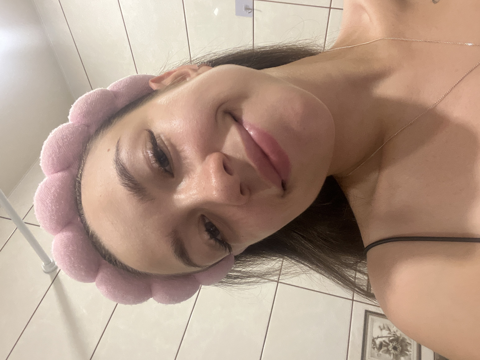 The image is a close-up selfie of a young woman indoors, likely in a bathroom. She is wearing a pink fluffy headband and a black spaghetti strap top. She has long dark hair and is smiling softly. Her skin appears clear and dewy, suggesting a skincare routine. The background shows tiled walls, a metal rod, and a framed picture on the left. The lighting is bright, possibly from overhead bathroom lights. There is no identifiable information suggesting the location is in Chaikovsky, Russia.