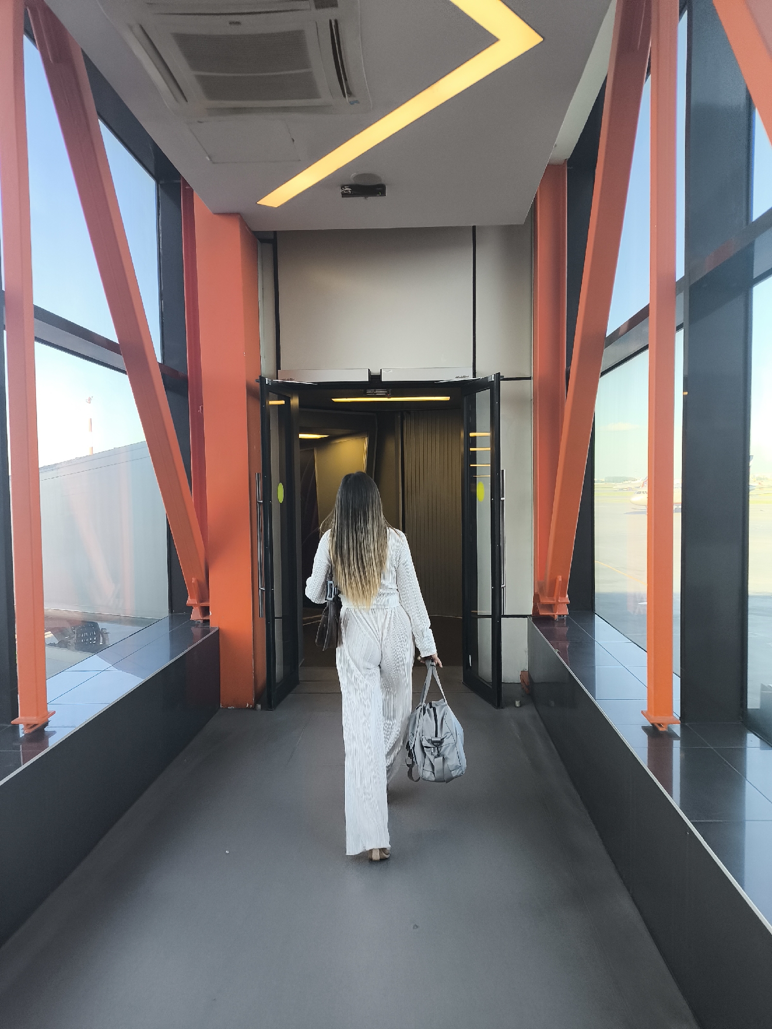 This image depicts the interior of an airport jet bridge or terminal walkway. A woman, viewed from behind, is walking away from the camera towards an open doorway at the far end. She has long, straight hair, dark at the roots and progressively lighter towards the ends. She is wearing a white or off-white textured jumpsuit or a matching two-piece garment, with light-colored footwear. She carries a dark brown shoulder bag on her left side and a light grey duffel bag in her right hand.
The walkway features a dark grey floor. Large, floor-to-ceiling glass windows line both sides, revealing a clear blue sky. Through the windows, an airport tarmac is visible; a commercial airplane's tail section is seen on the right, and a white building with a distant red tower is visible on the left. Bright orange, angular structural beams are prominently integrated into the window frames. A low, black-surfaced ledge runs along the base of the windows. The ceiling is light grey, equipped with an overhead air conditioning unit and a distinctive yellow, angular LED strip light. The open doorway leads into a darker internal area.
Location: Airport terminal, Unknown City, Unknown Country.