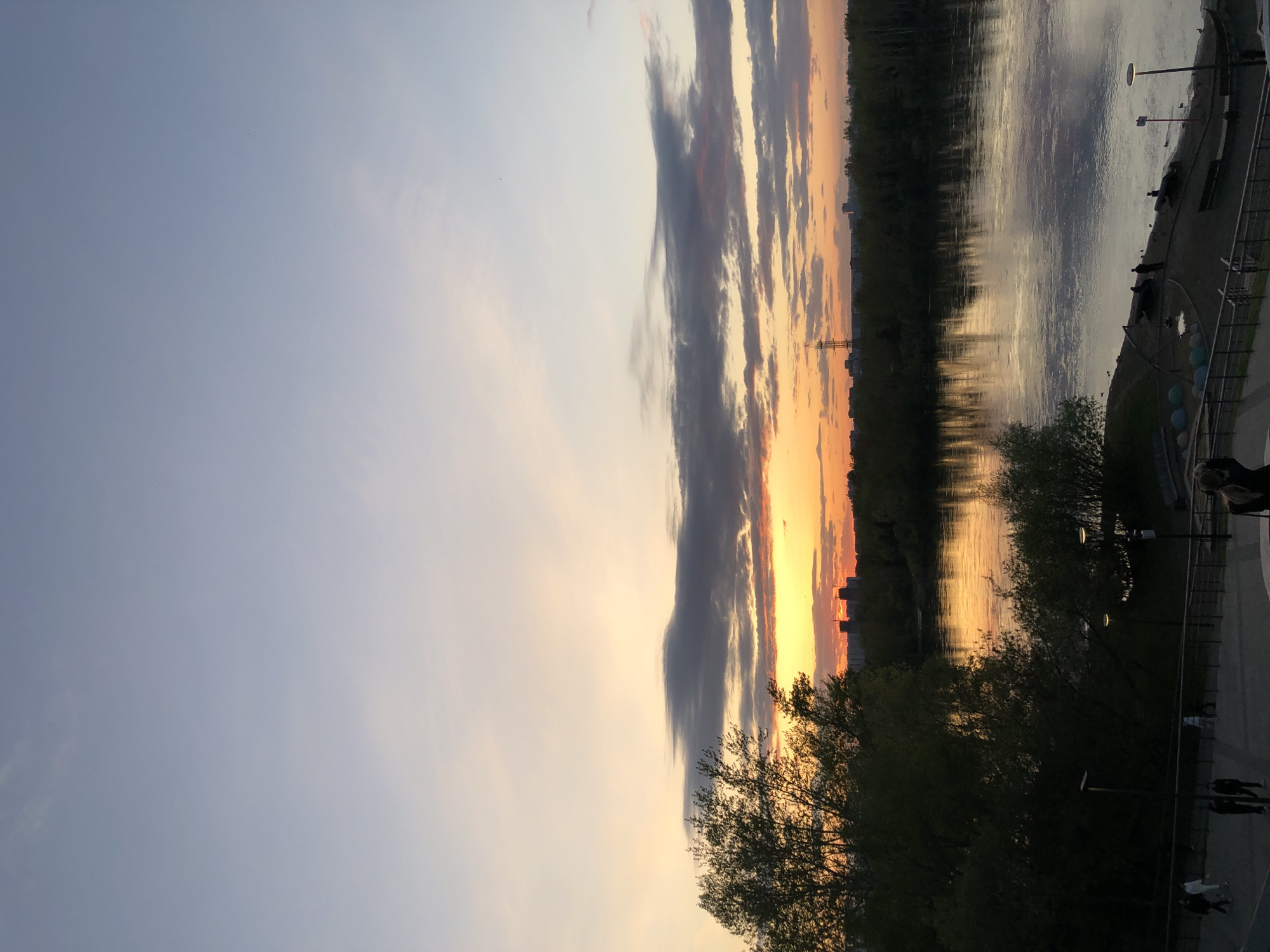 The image depicts a sunset over the Yenisey River in Krasnoyarsk, Russia. The sky transitions from light blue to orange with striated clouds. The river reflects the orange hues. A line of trees and buildings marks the riverbank. A pedestrian walkway with streetlights is visible in the foreground, with people walking along it. A large tree partially obscures the view in the lower portion of the image. A transmission tower is visible in the midground on the opposite bank.