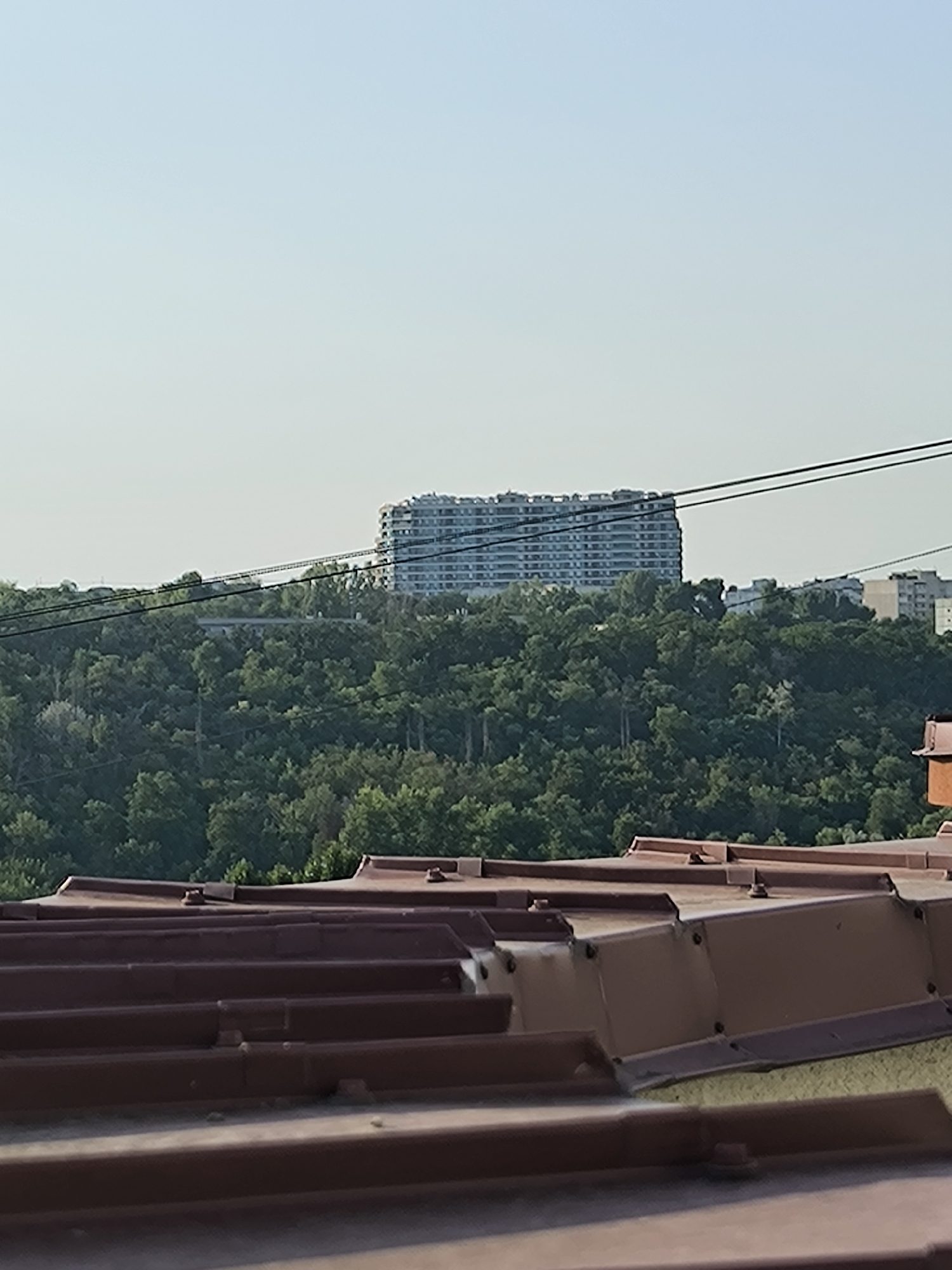 This is a photograph taken from a rooftop in Chișinău, Moldova. In the foreground, a close-up view of a brown corrugated metal roof is visible, with several raised seams and visible fasteners. The roof appears to be part of a building. In the mid-ground, a dense green forest covers a hill. Beyond the forest, a long, multi-story white building with many windows is situated on a higher elevation. The building has a horizontal architectural style, suggesting it could be an apartment complex or a hotel. Two dark, thin power lines stretch horizontally across the image in front of the distant building and above the forest. The sky is a clear, pale blue, indicating it is daytime. No people are visible in the frame.