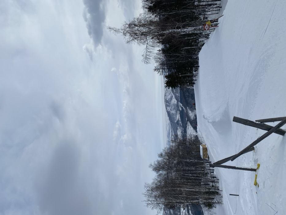 The image displays a snow-covered ski slope within a mountainous landscape in Novoabzakovo, Russia, under an overcast, cloudy sky. The foreground presents a wide, groomed ski path. To the immediate left, several wooden posts forming a broken barrier are partially buried in the snow, with a yellow object visible near their base. Along the left perimeter of the slope, a dense line of deciduous trees with bare branches and dark coniferous trees are visible. Further down this left edge, a small, light-colored building with a dark roof is situated among the trees. In the mid-ground, a prominent snow ramp or jump is sculpted into the slope. A single individual, appearing as a small red figure, is located on the slope past this ramp. The right side of the slope is also bordered by a mix of bare deciduous and coniferous trees. A yellow and red sign, featuring the number "4", is affixed to a pole or tree on this right side. The background consists of multiple layers of snow-covered mountains and hills extending to the horizon under the consistent grey sky.