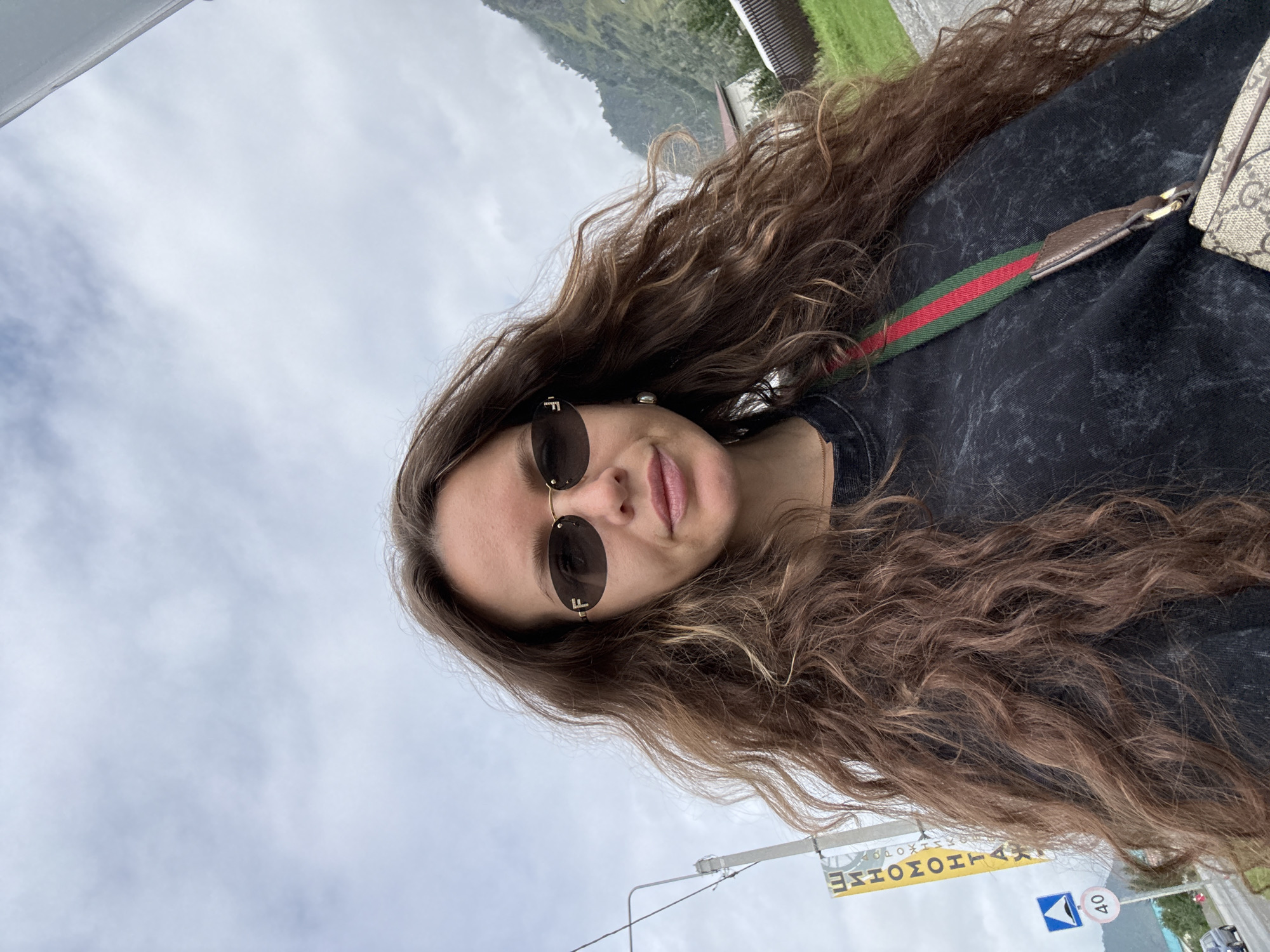 In this selfie, a woman with long, wavy brown hair and sunglasses stands outdoors. She is wearing a dark, possibly distressed, t-shirt and a crossbody bag with a prominent green and red stripe and a beige patterned material. A road sign with the number "40" is visible in the background, along with a yellow sign with Cyrillic text that reads "ШИНОМОНТАЖ", indicating a tire fitting service. Mountains and some buildings are visible further in the background under a cloudy sky. The scene is located on a road in the city of Chemal, Russia, indicated by the specific signage and the mountainous terrain typical of the Altai region.