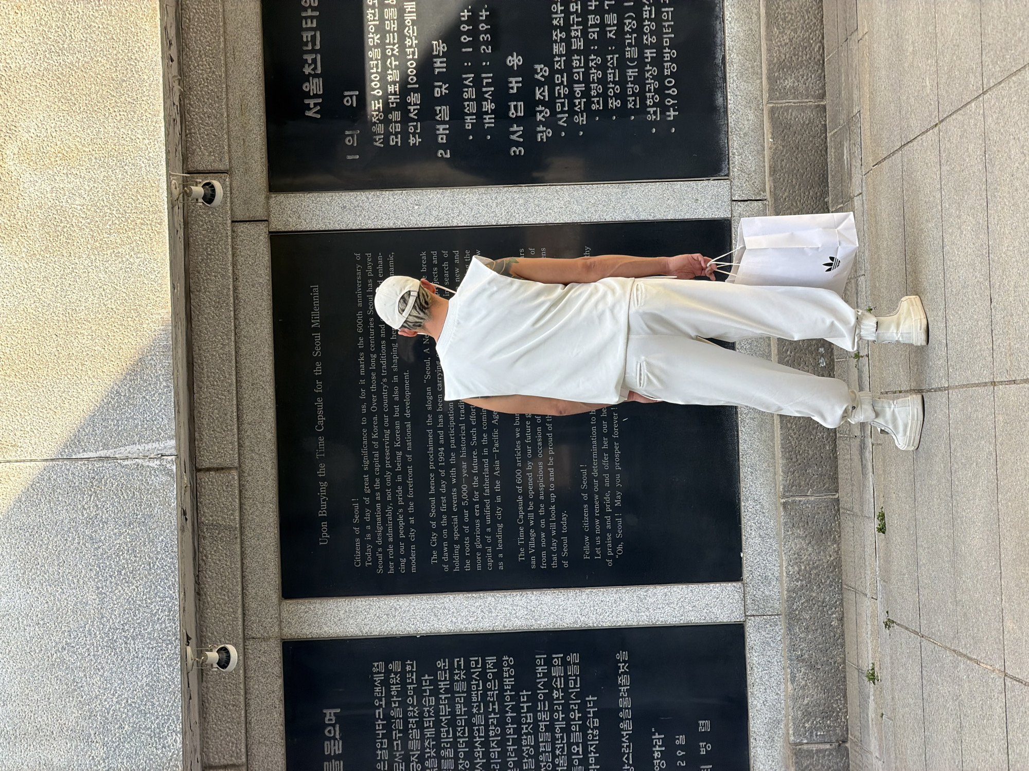 A person in all-white clothing, including a hat, carrying a white shopping bag with an Adidas logo, stands in front of a memorial or commemorative wall in Seoul, South Korea. The wall consists of multiple dark stone panels with inscriptions in both Korean and English. The panels discuss the history of Seoul, its designation as the capital, and the burying of a time capsule. The person appears to be viewing or reading the inscriptions. The scene is outdoors on a paved walkway or sidewalk. There is a security camera mounted on the wall.
