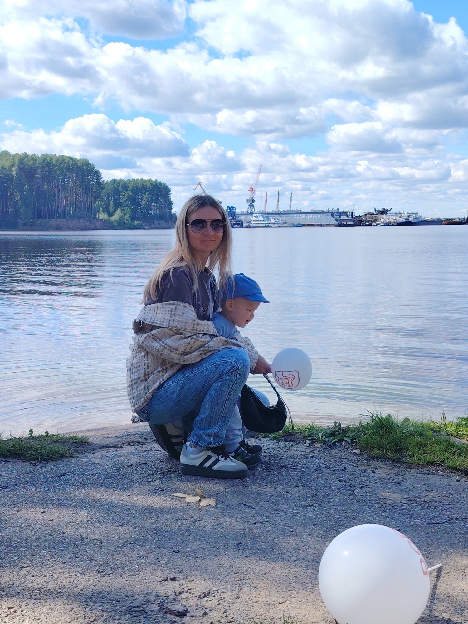 A woman with blonde hair and sunglasses is crouching by a body of water, with a child wearing a blue hat in her lap. The woman is wearing a plaid jacket over a dark t-shirt and blue jeans. The child is wearing a blue shirt. They are on a gravelly shore with grass in the background. In the foreground, there is a white balloon. In the background, there is a wide river with a forested shoreline on the left, and industrial structures and ships on the water in the distance, which are part of the Chaikovsky port area. The sky is blue with fluffy white clouds.