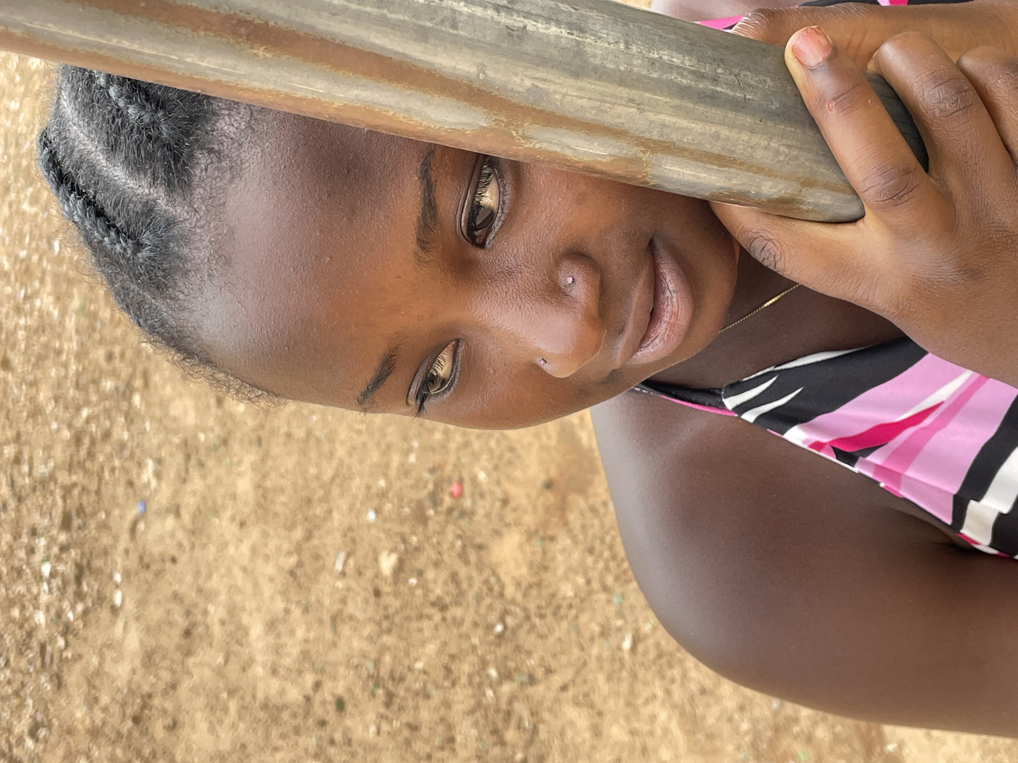 A close-up, angled portrait captures a young person, likely female, with dark skin, looking directly at the viewer with a subtle smile. Her head is tilted, resting against a thick, weathered, cylindrical pole or beam that runs diagonally across the top of the frame. Her dark eyes reflect a bright light source, and she has a small, light-colored stud in her right nostril. Her hair is styled in visible braids (cornrows) on the side of her head. She is wearing a top or dress with a distinct pattern of black, pink, and white stripes, and a slender gold chain is visible around her neck. Her hands are gripping the pole, which appears to be made of wood or aged metal. The background is an out-of-focus expanse of sandy or dusty ground, characteristic of an outdoor setting. The bright, natural lighting suggests it is daytime. The image conveys a sense of calm and direct engagement, set in what appears to be Barakin Akawo, Nigeria.