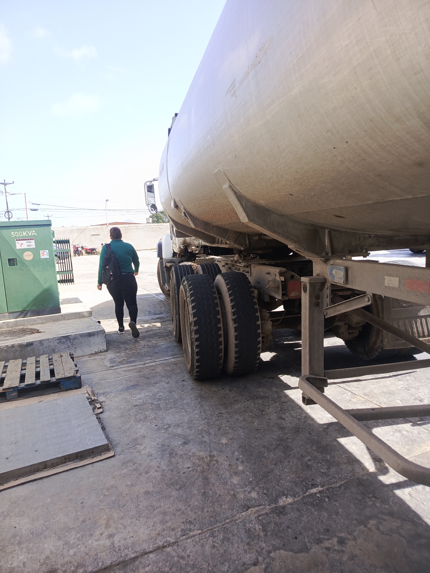The image shows an industrial area in Juan Griego, Venezuela. A woman with a dark green shirt and black pants walks away from the camera, past a large tanker truck. A green electrical box is visible to the left, with a wooden pallet and concrete slabs nearby. The sky is overcast, and the pavement is concrete.