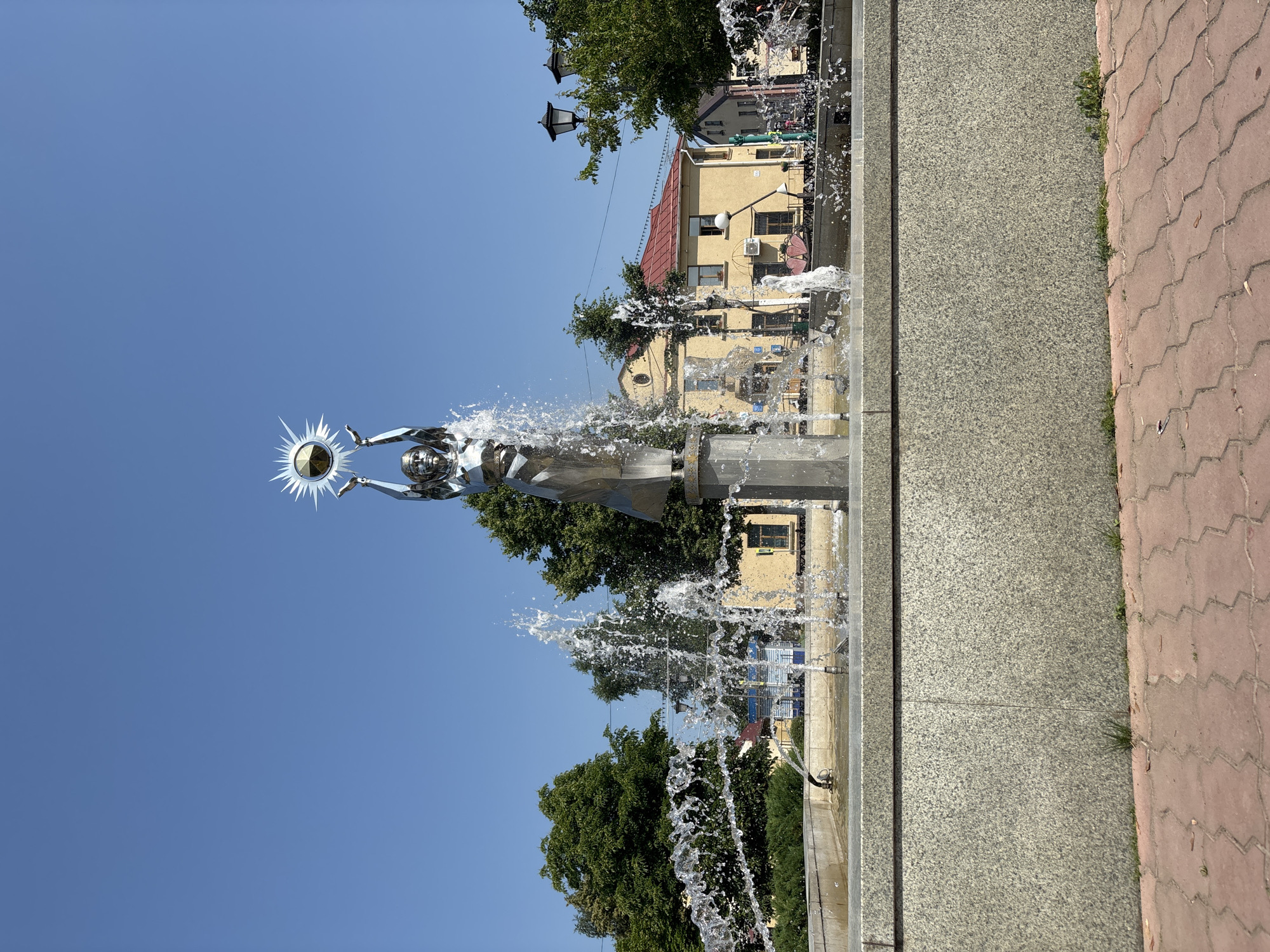 A statue of a woman holding a sun-like object stands in a fountain in Pastavy, Belarus. The statue is made of polished metal and the sun-like object has rays emanating from it. Water sprays from the fountain around the statue, creating a mist. In the background, there are buildings and trees. The sky is clear and blue. The statue is likely a symbol of the sun or light.