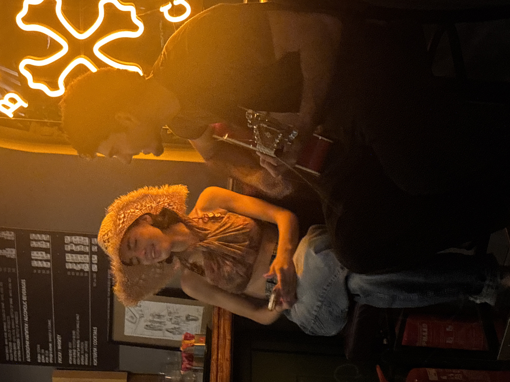 In Sofia, Bulgaria, a woman in a large straw hat and a patterned halter top sits with a phone in her hands, while a man plays an acoustic guitar. They are seated in a dimly lit establishment, possibly a bar or cafe, with a menu board visible behind the woman listing alcoholic beverages and cocktails. A bright neon light fixture, shaped like a four-leaf clover with a central swirl, illuminates the background with a warm, orange glow. The woman appears to be smiling, her eyes closed, as if enjoying the music or the moment. The man is focused on his guitar, his profile visible against the neon light. The overall atmosphere is intimate and relaxed, suggesting a live music performance or a casual gathering.