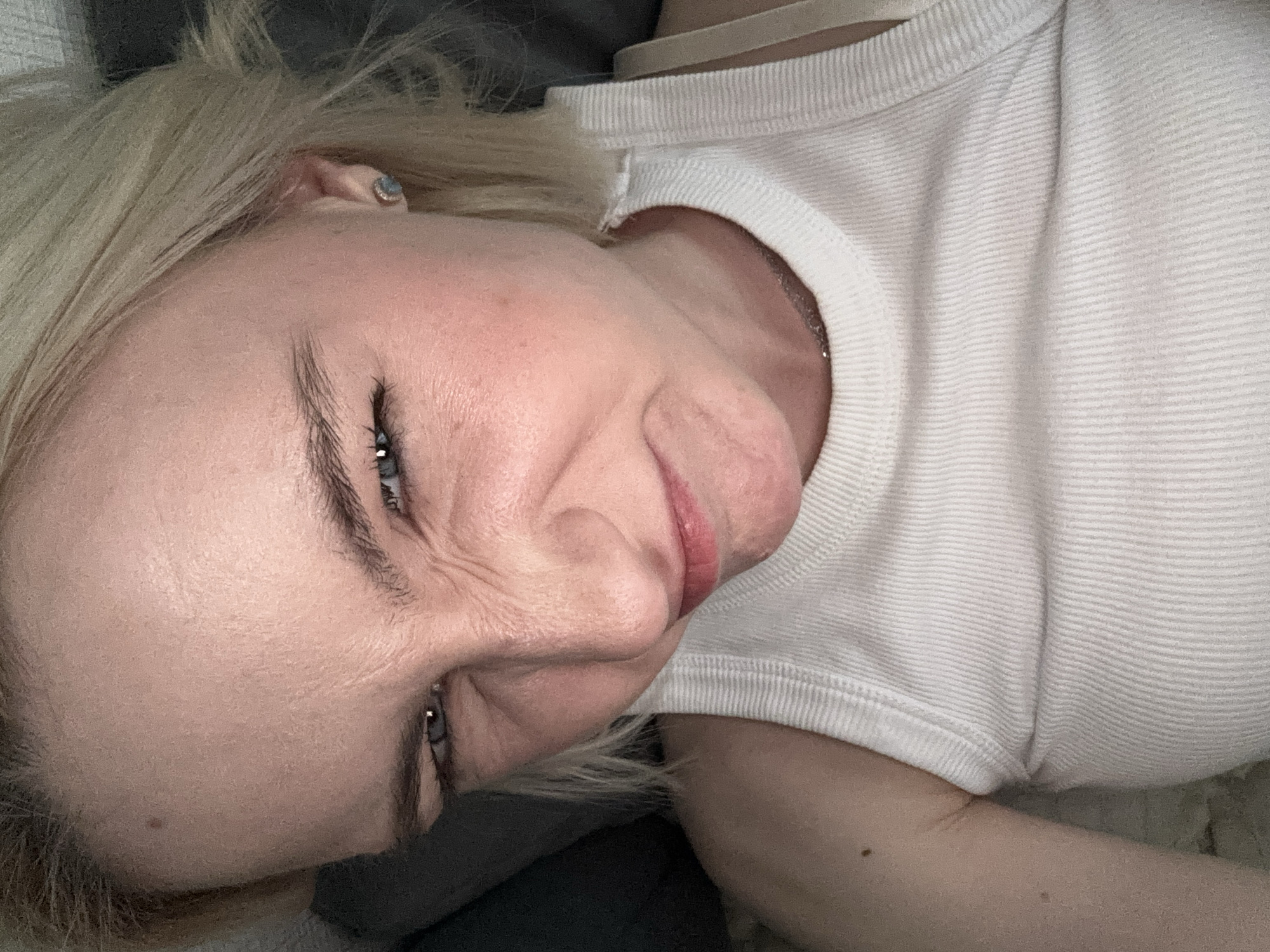 A blonde woman in a white ribbed top is lying down, possibly on a bed, indoors. Her face is angled towards the camera with a slight smile. She appears to have an earring in her left ear. No location in Vsevolozhsk, Russia is discernible in this image.